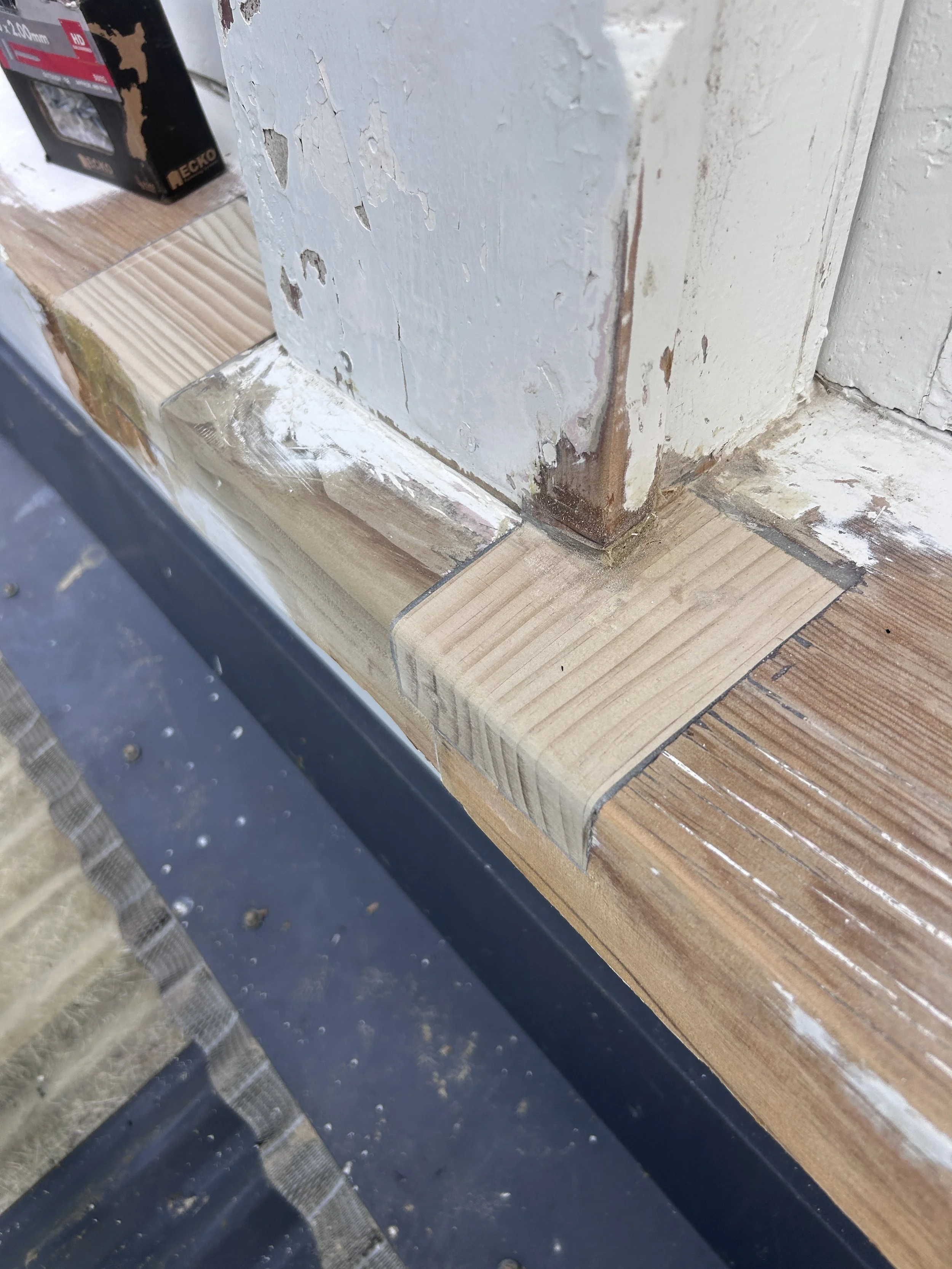 Close-up photo of a corner of a wooden surface with peeling white paint and some rusted metal at the edge, with a black box in the background.
