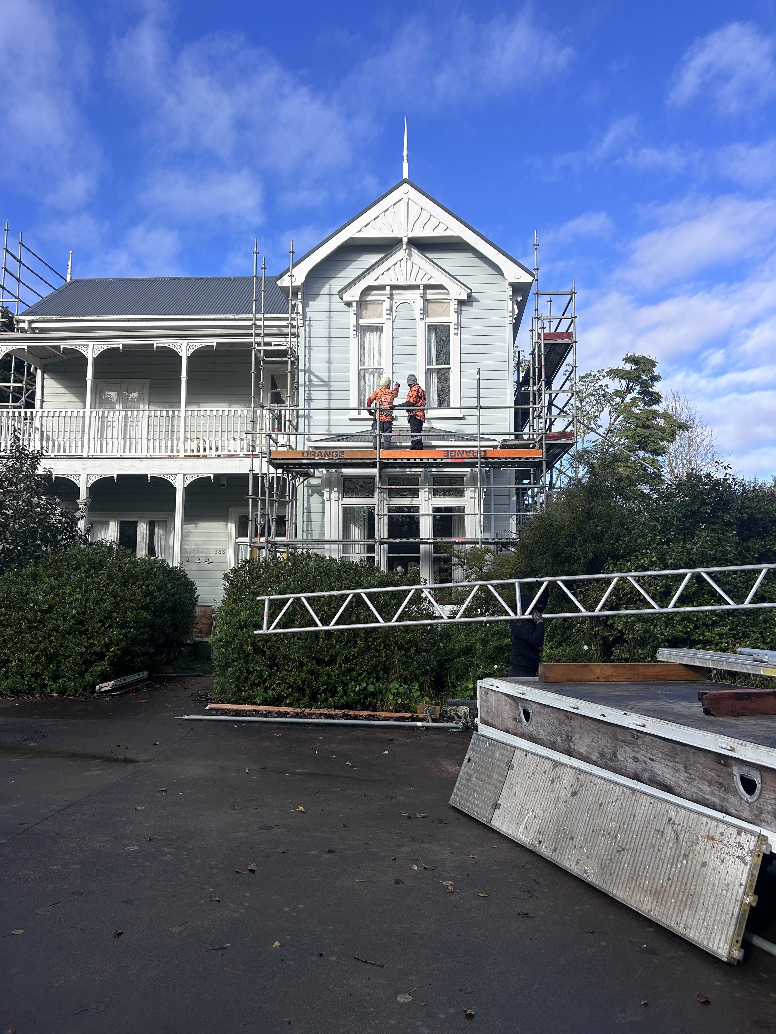 People working on scaffolding outside a light blue Victorian house with white trim, surrounded by greenery, under a bright blue sky with some clouds. Construction equipment and ladders are present.