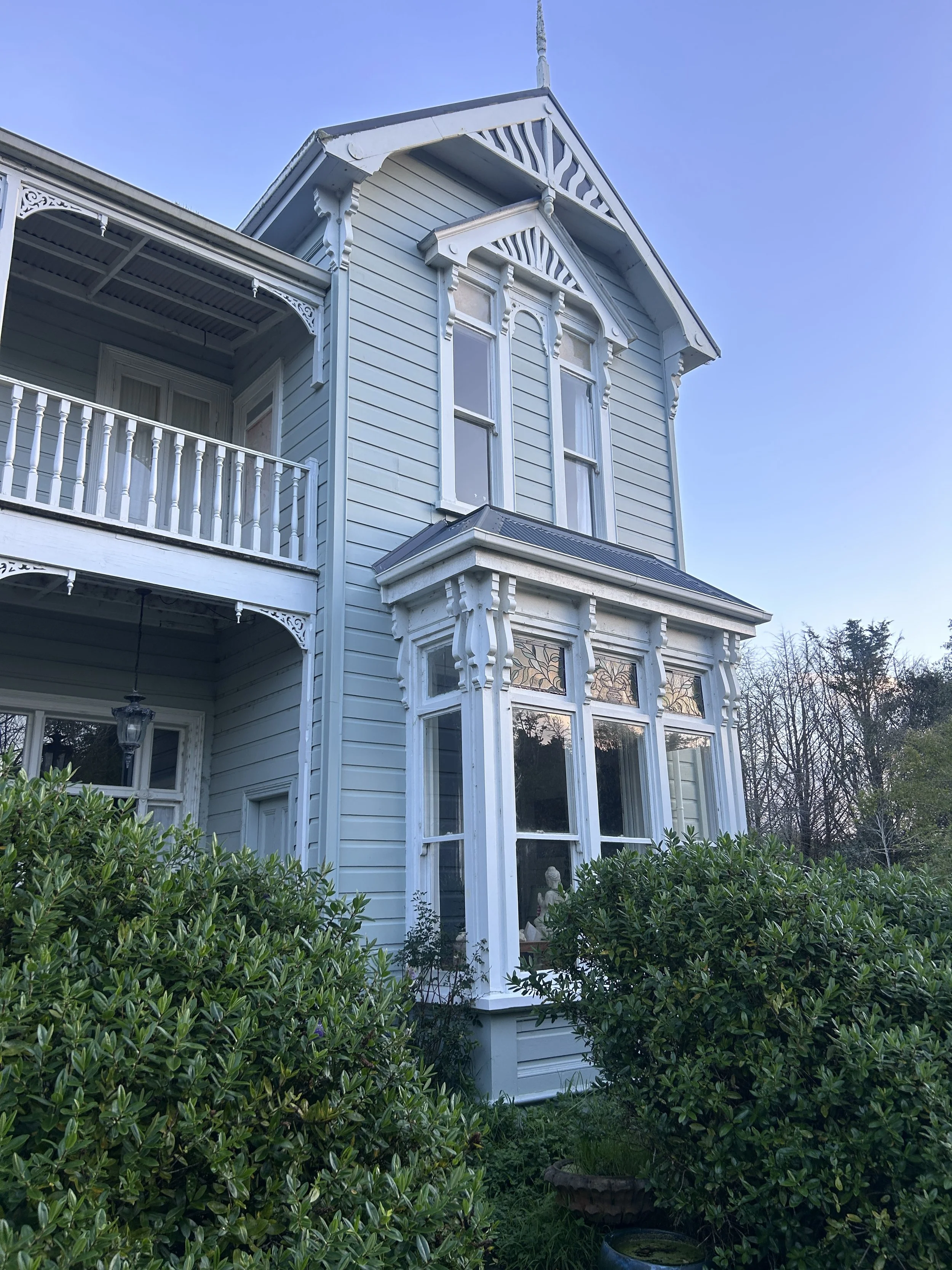 A white Victorian-style house with decorative trim, large windows, and a balcony surrounded by lush green bushes and trees.