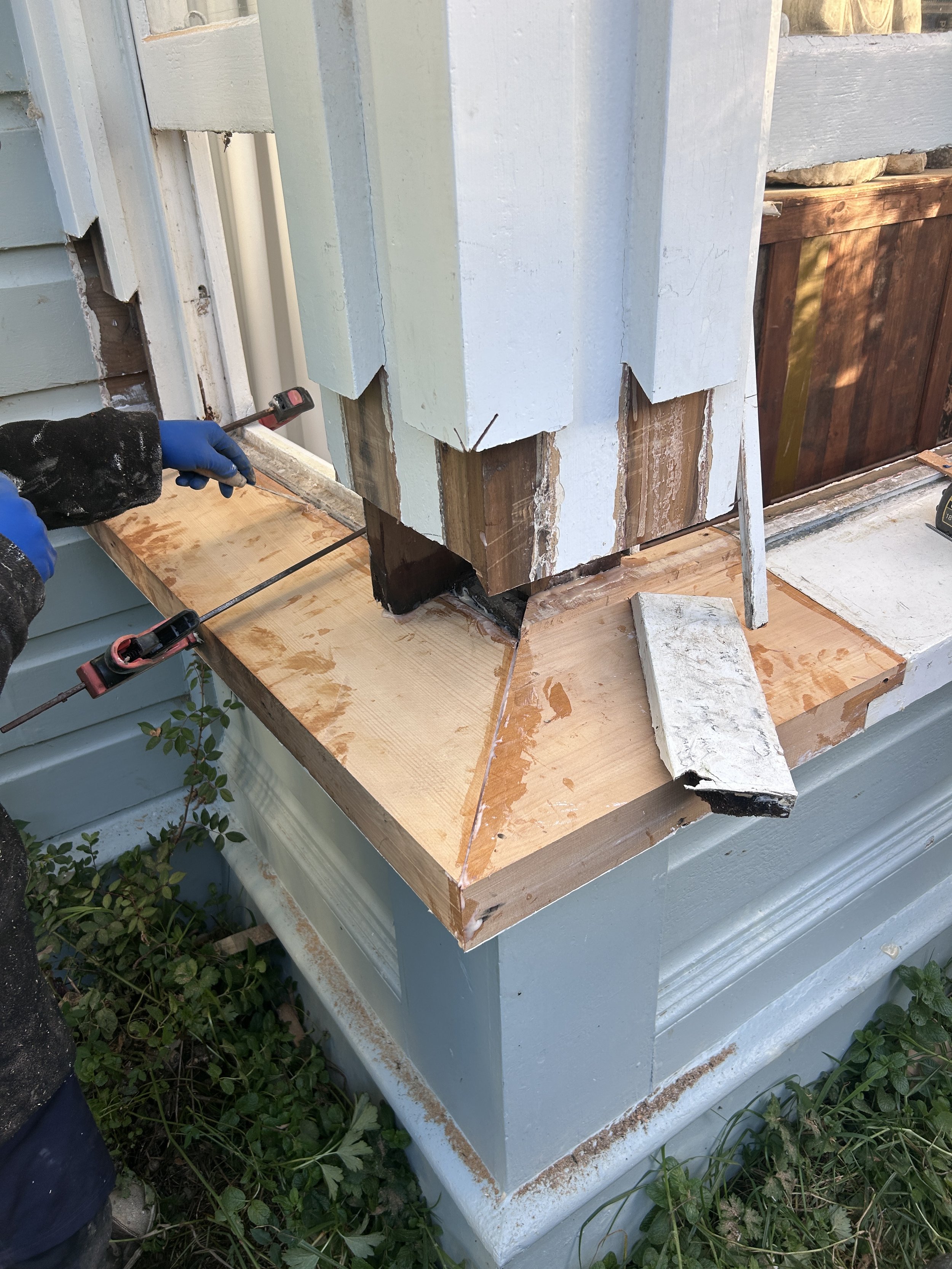 Someone repairing or inspecting a house corner with wooden trim, clamps, and paint on the siding, outdoors with plants nearby.