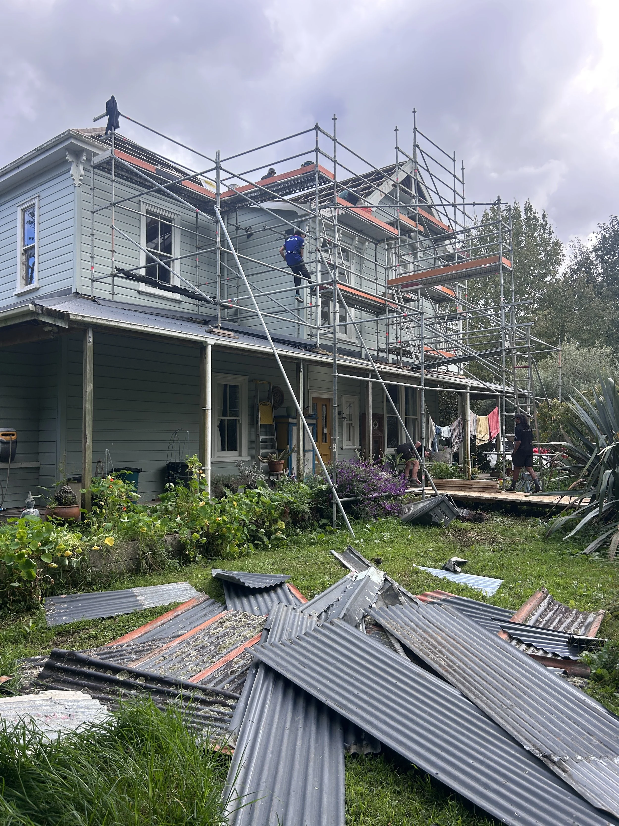 House with scaffolding around it during construction or repair, with debris and damaged metal roofing sheets scattered on the yard.