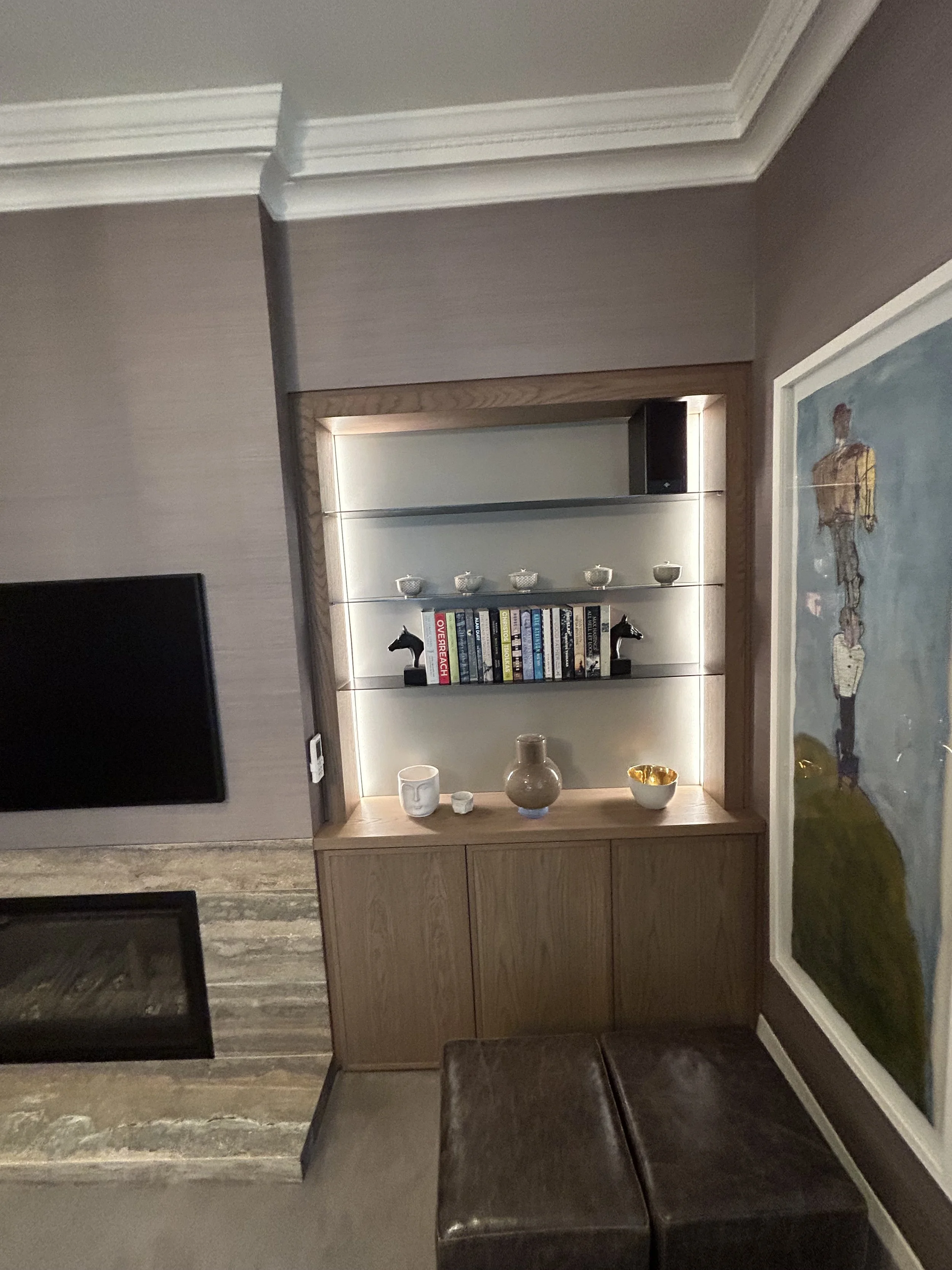 A corner of a room showing a wooden display shelf with decorative bowls and books, a framed painting of a person, a television, and two black horse head sculptures, with flooring and wall molding visible.
