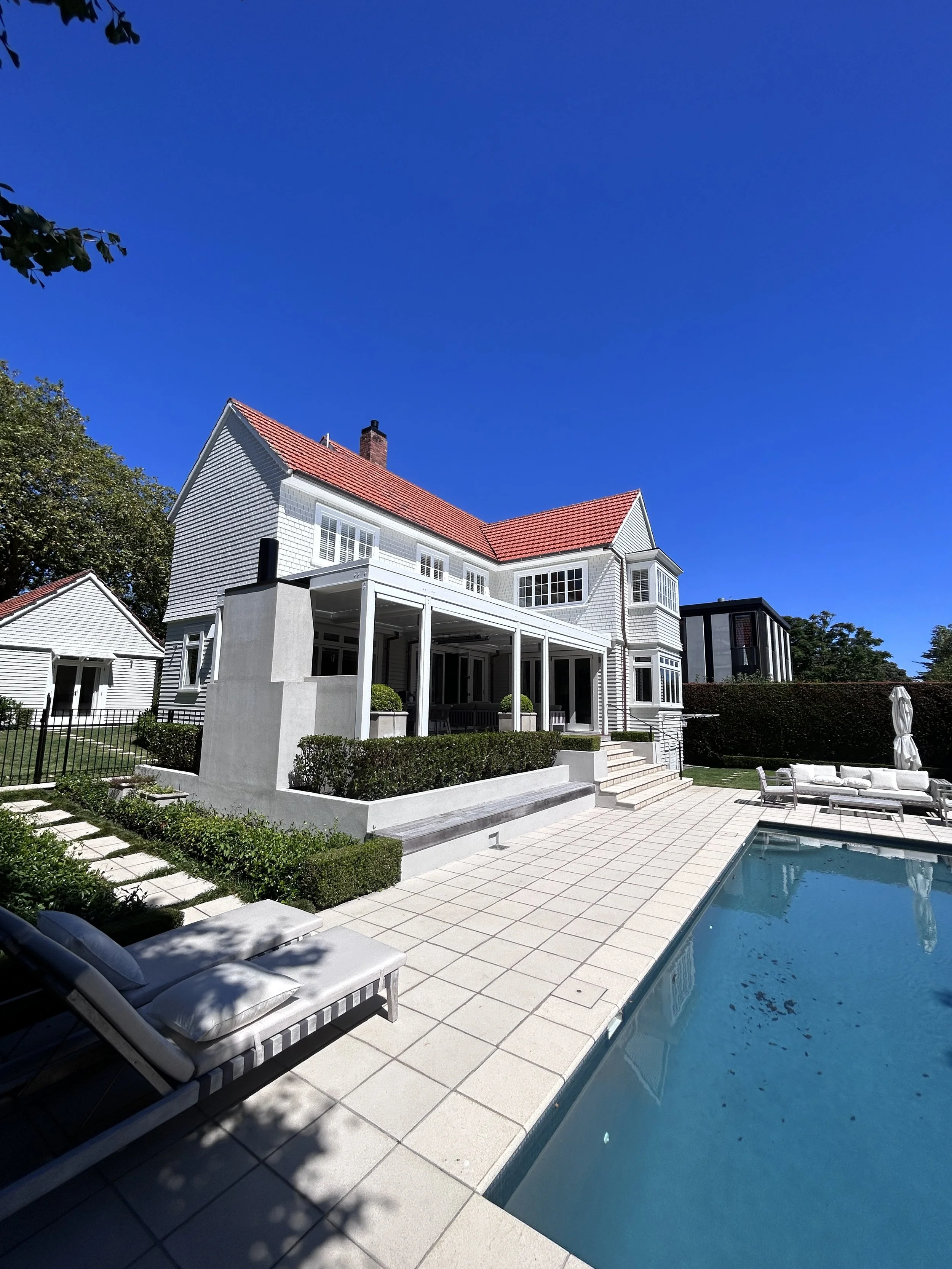 A large white house with a red roof, a swimming pool, and outdoor seating area under a clear blue sky.