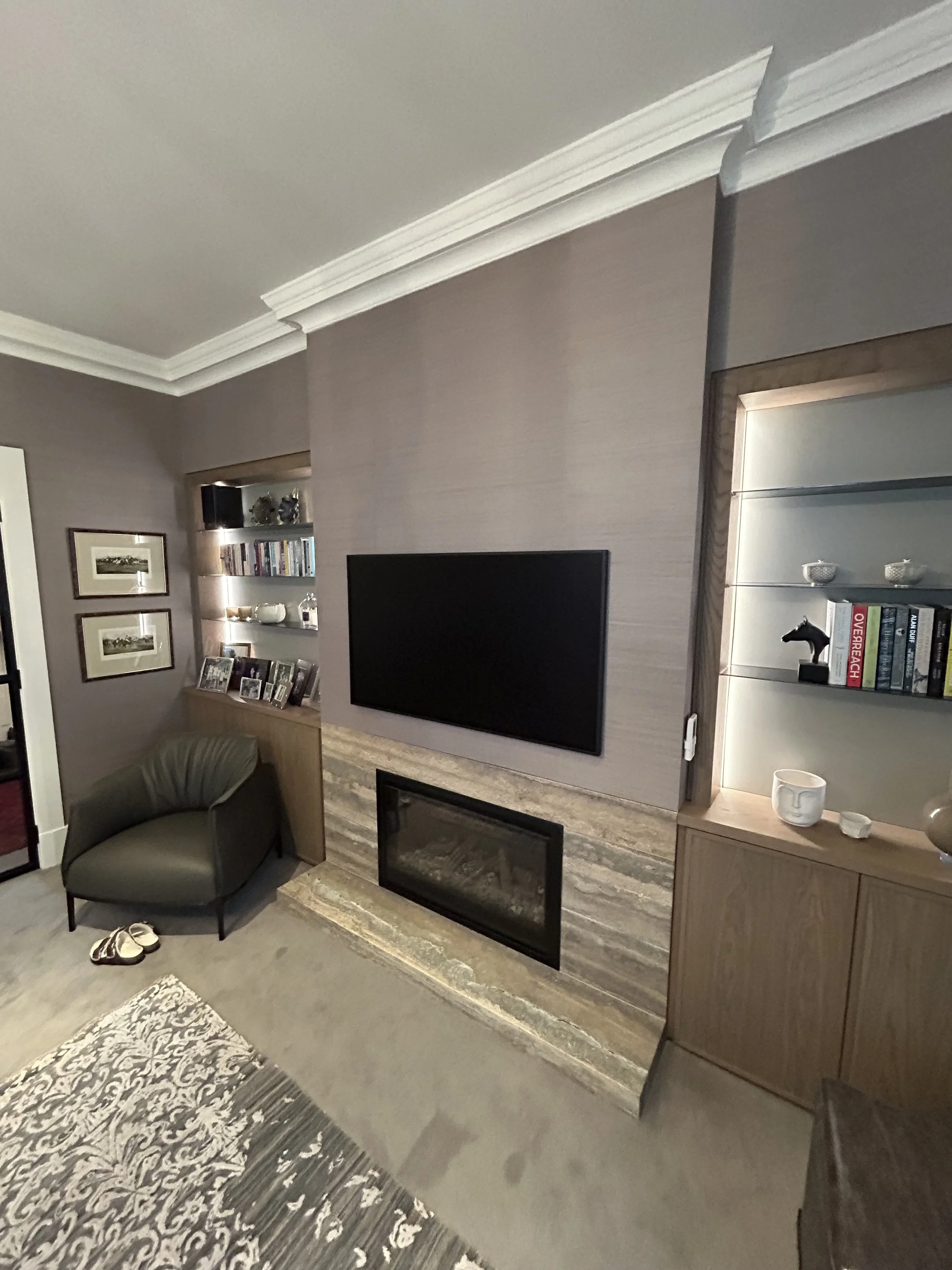 Living room with a flat-screen TV, framed pictures on the wall, bookshelves, a gray armchair, a fireplace, and a patterned rug.
