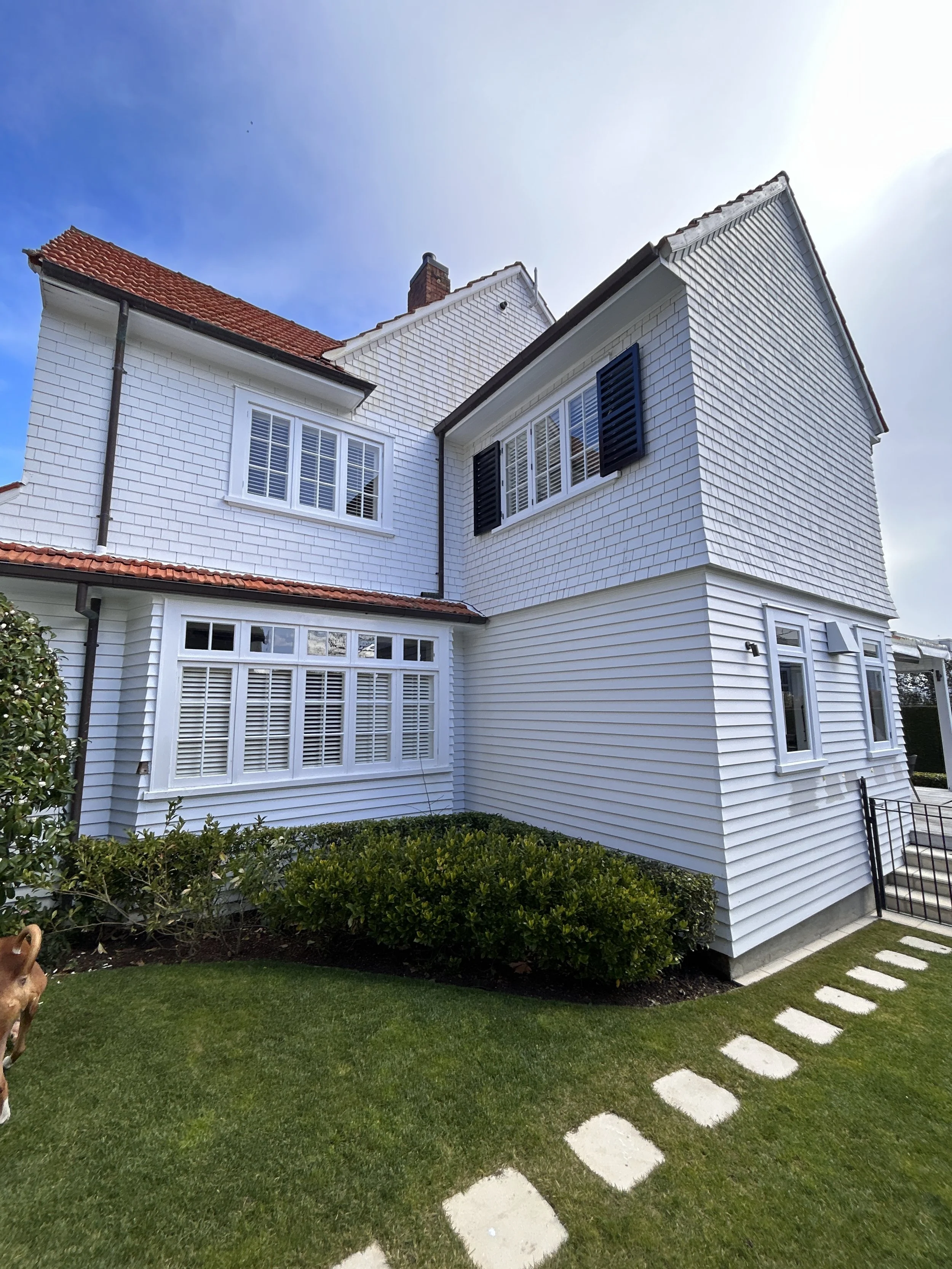 White two-story house with red tile roof, black shutters, large windows with white frames, a brick chimney, and a small front garden with trimmed green bushes. Stone pathway leads to the entrance on the right.