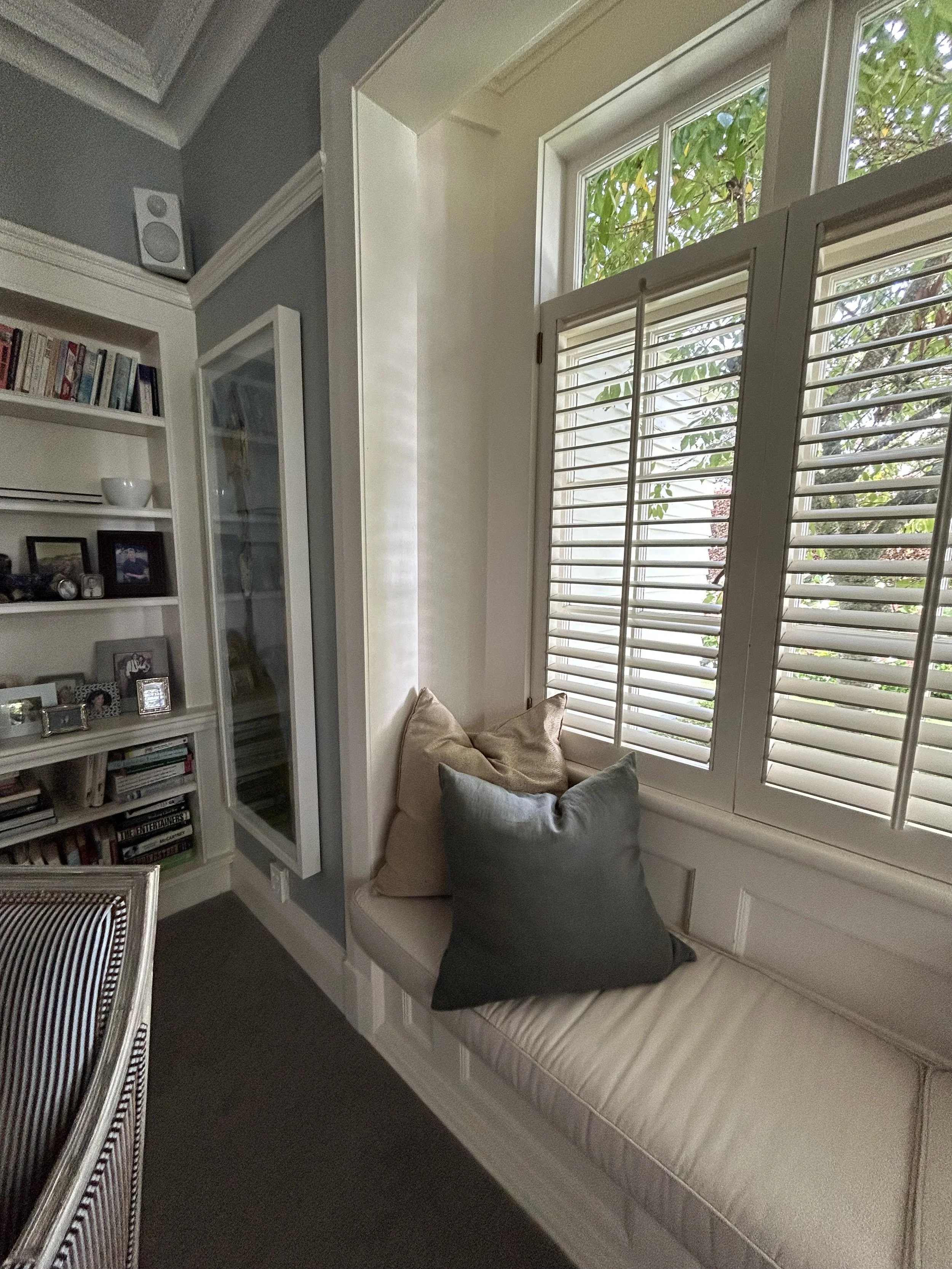 A cozy window nook with a cushioned bench and a group of pillows, next to a large window with white shutters outside view of green leaves.