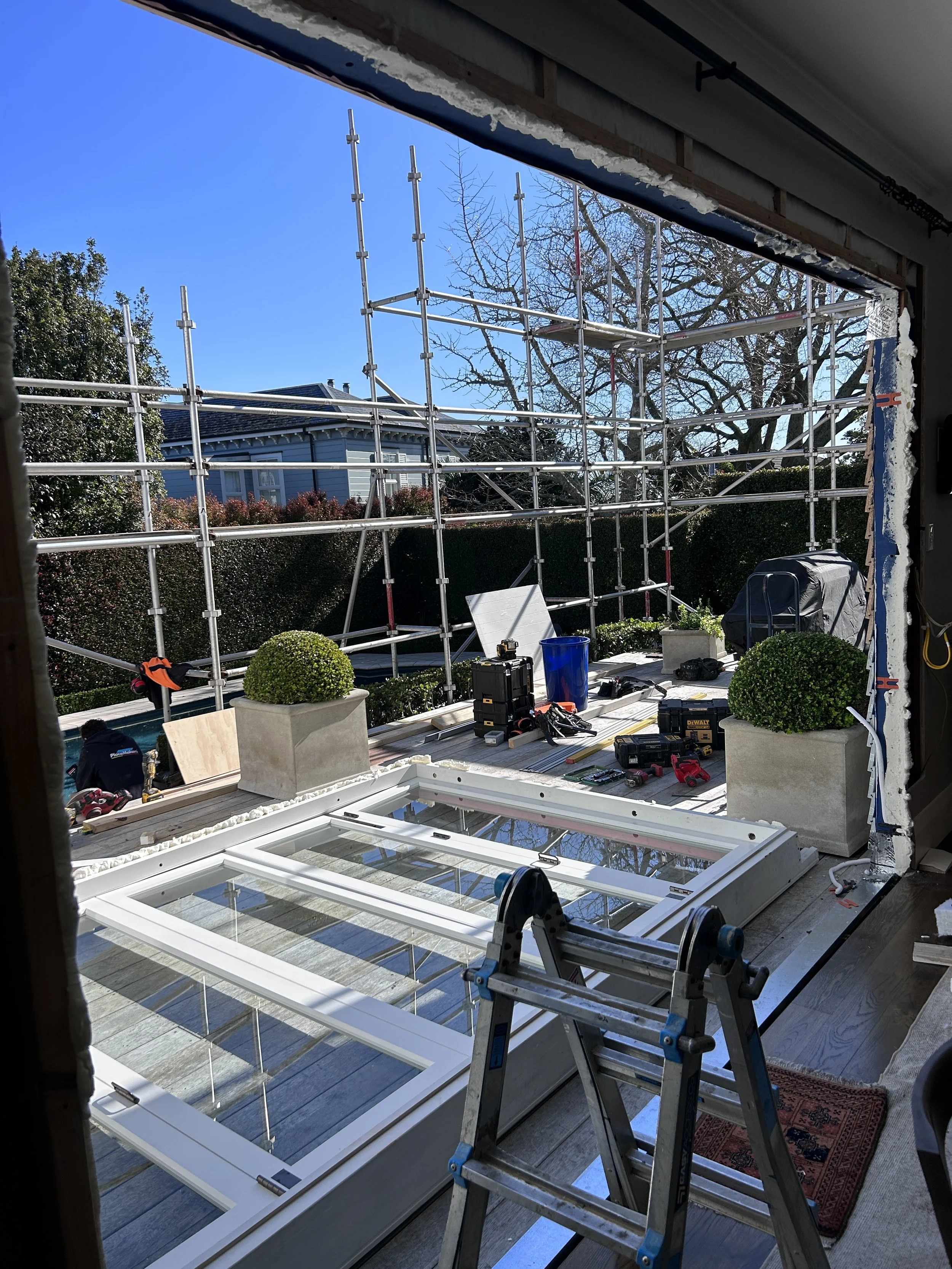 View of a house under construction with scaffolding outside, a frosted window on the deck, and various construction tools and equipment scattered around.