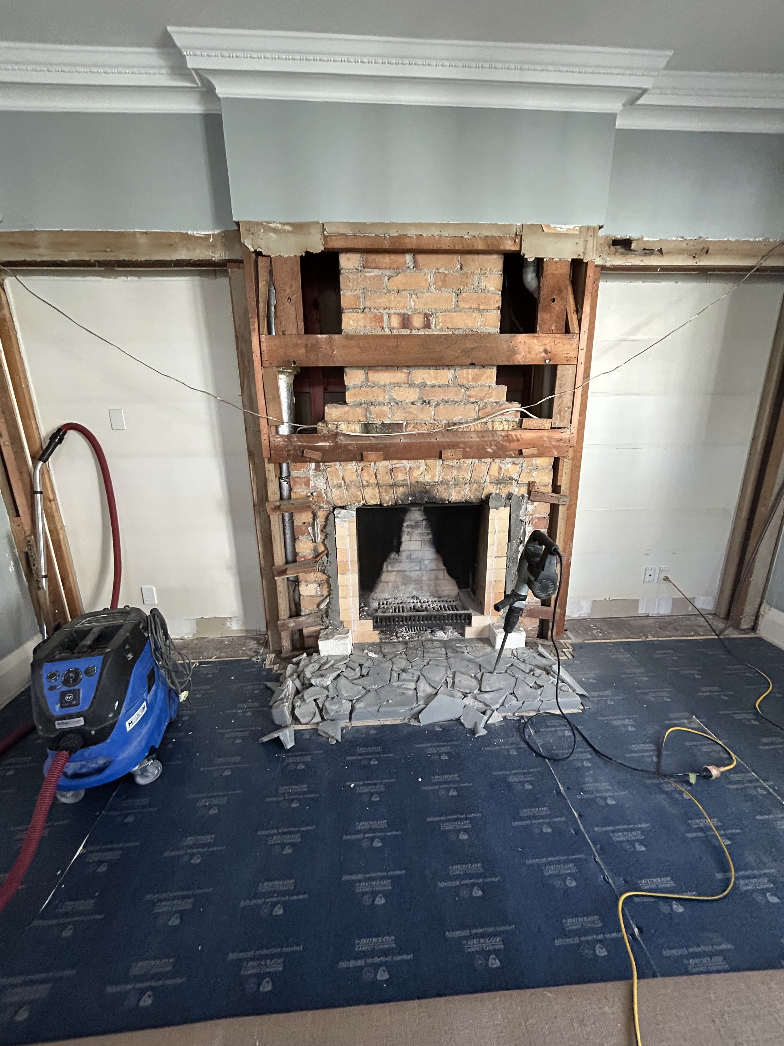 Living room fireplace undergoing renovation, with exposed brick and wooden framing, tools, and construction materials on the floor.