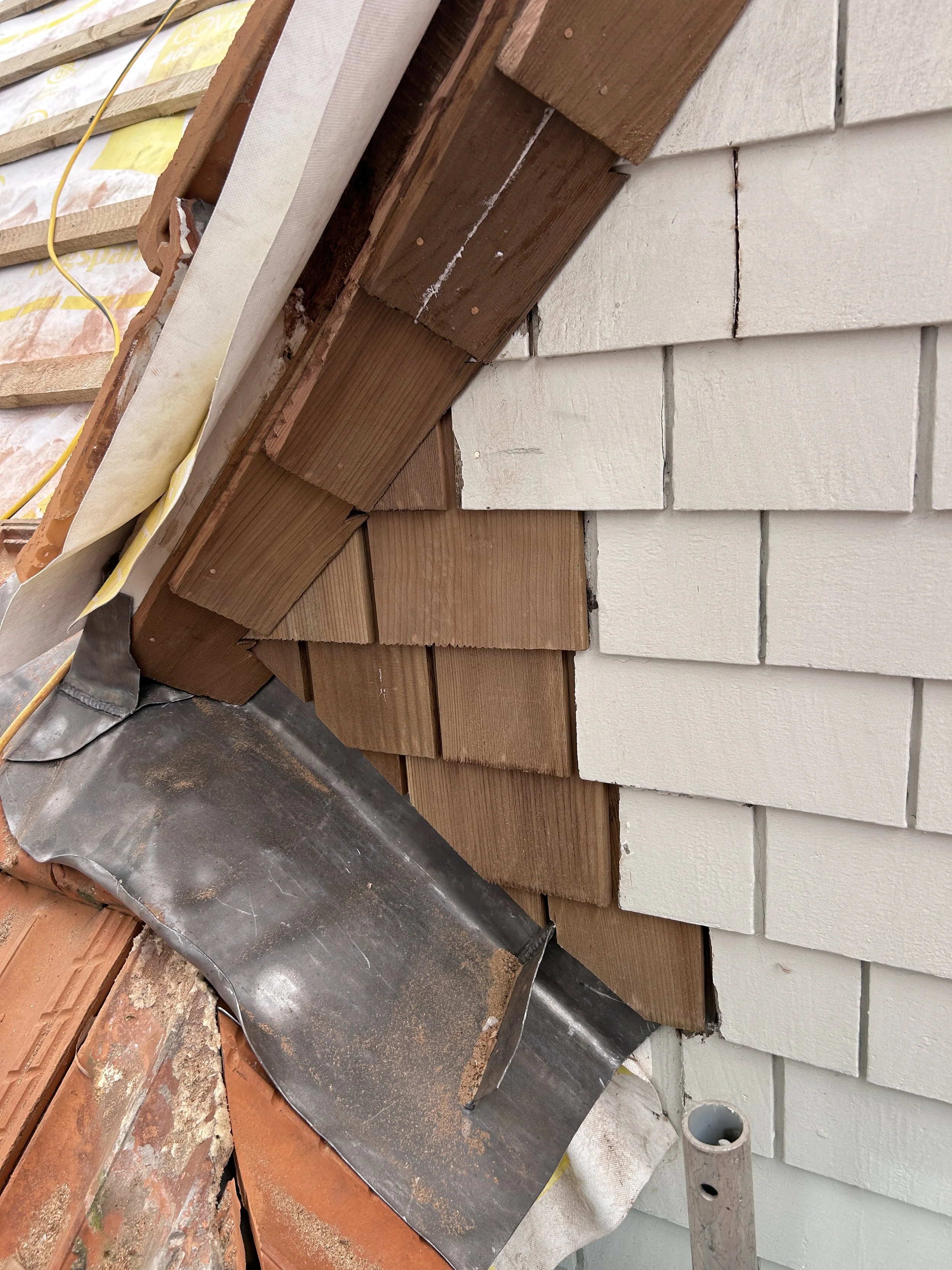 Close-up of a section of a house's exterior wall under construction, showing white siding, brown wooden trim, and part of a metal roofing edge. The image also depicts construction materials and tools.