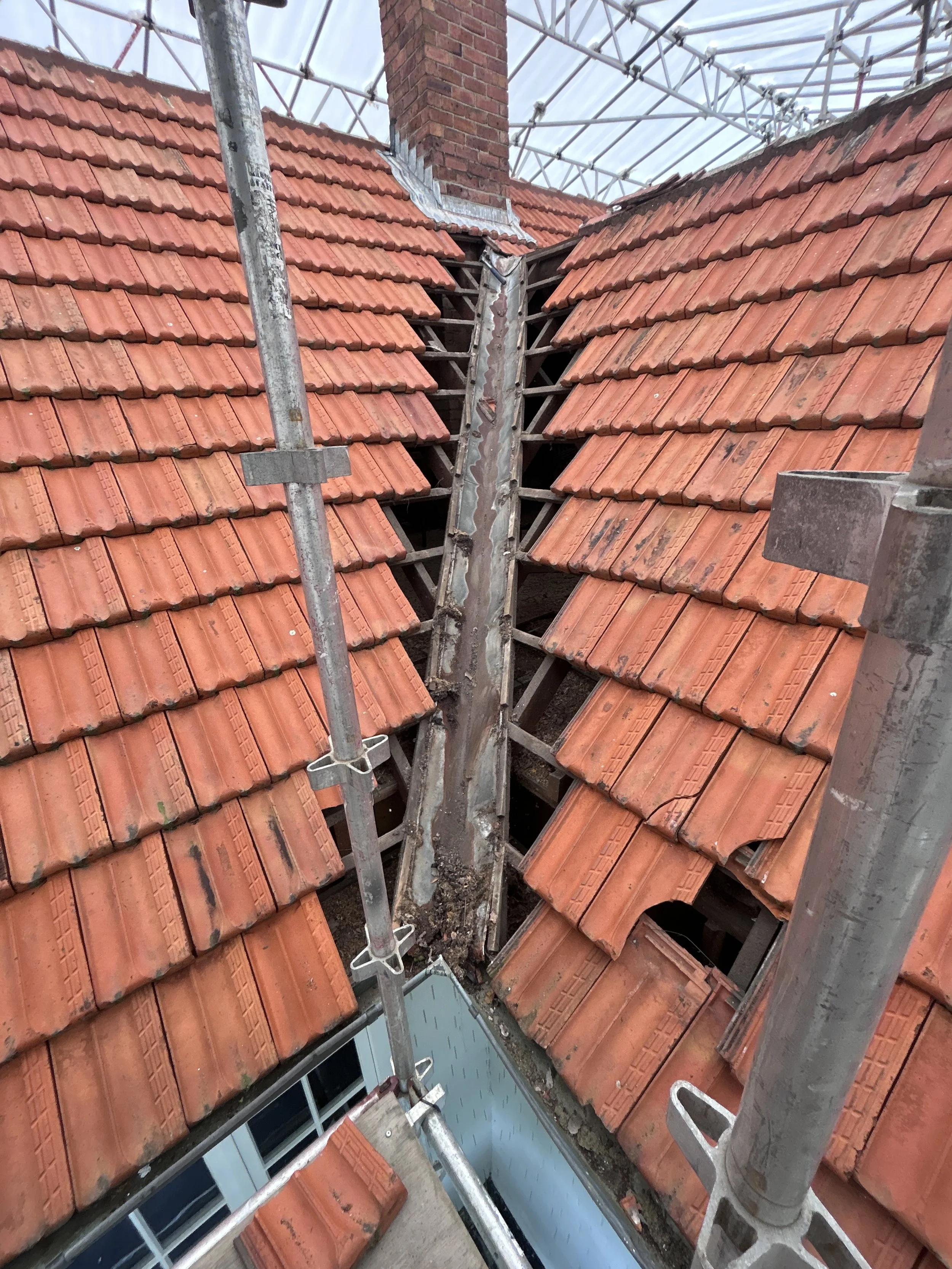 View of a roof under construction with orange clay tiles, scaffolding in place, and a chimney. Some roof tiles are missing, revealing wooden beams underneath.