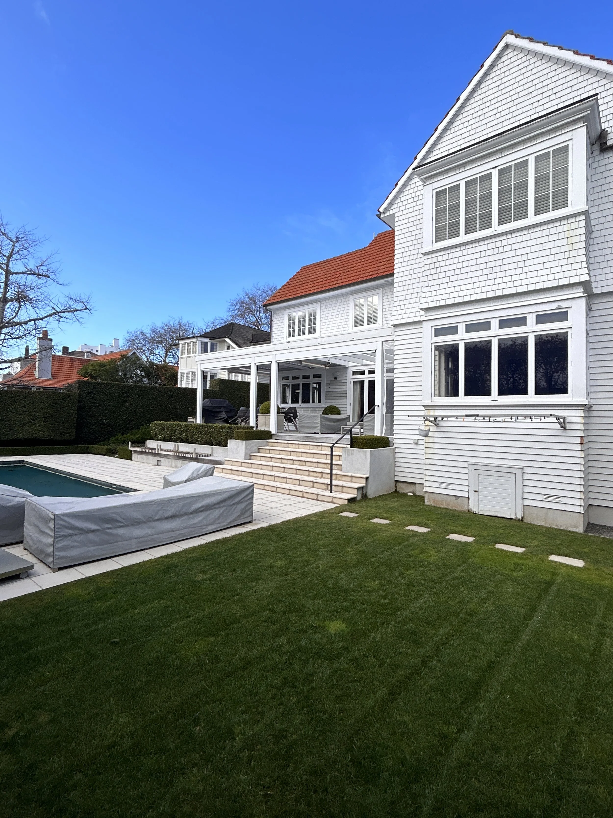 Backyard with green lawn, patio area with outdoor furniture covered with gray protective covers, white house with large windows, stairs, and a covered porch, under a clear blue sky.