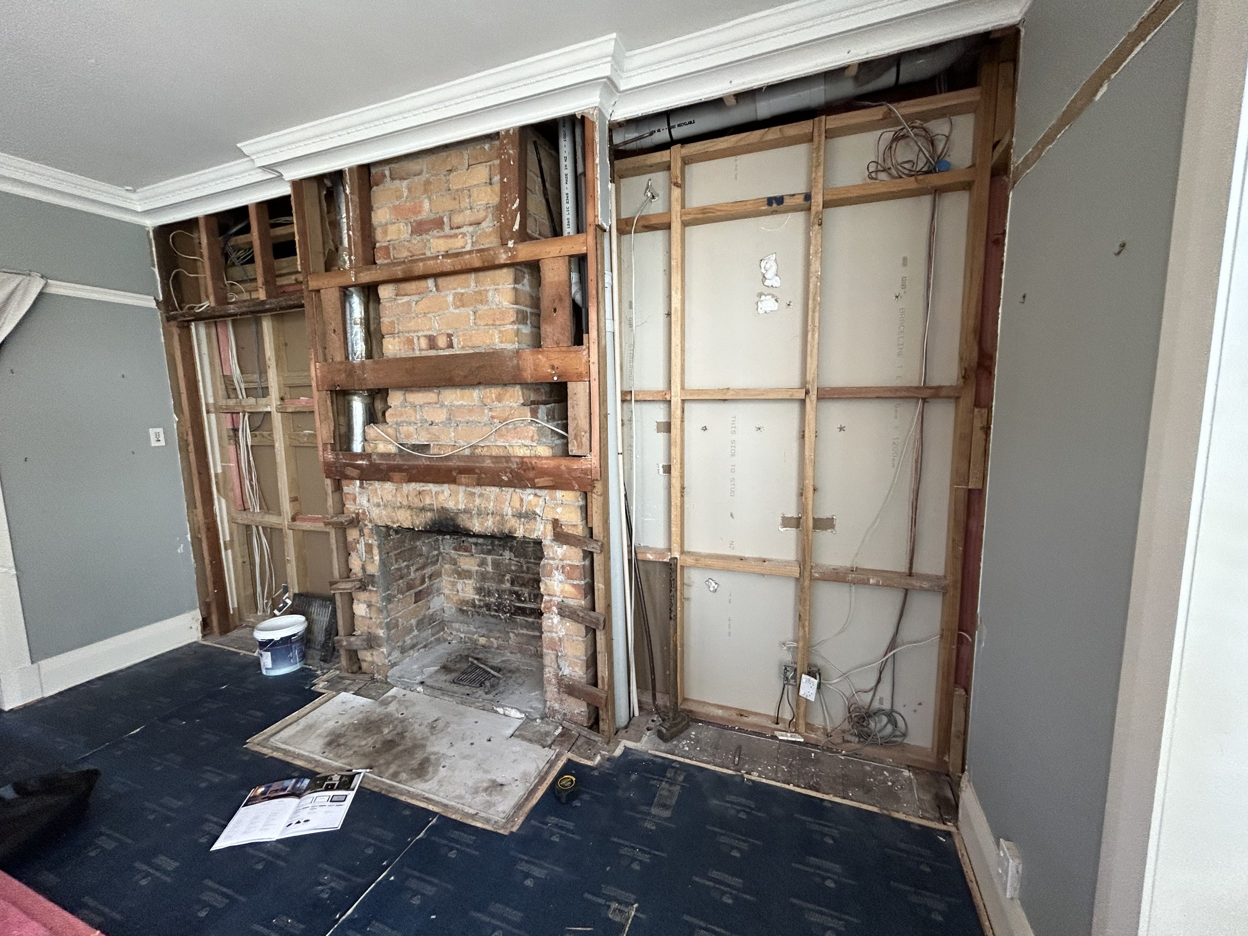 Interior room under renovation with exposed wooden framing and a brick fireplace, construction tools and materials scattered on the floor.