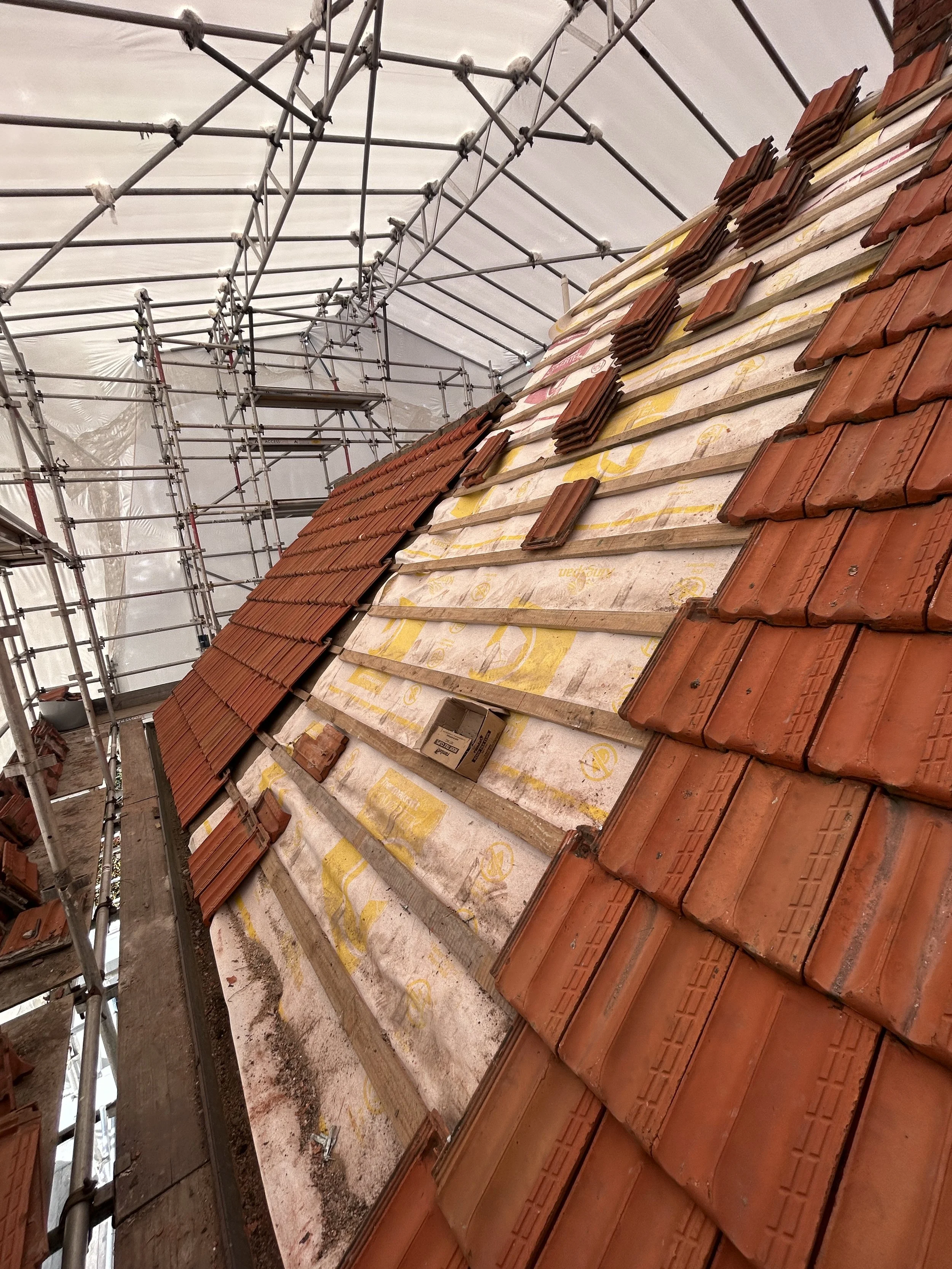 Roof under construction with red clay roof tiles being installed on wooden battens, metal scaffolding surrounding the building, and a white protective sheet overhead.