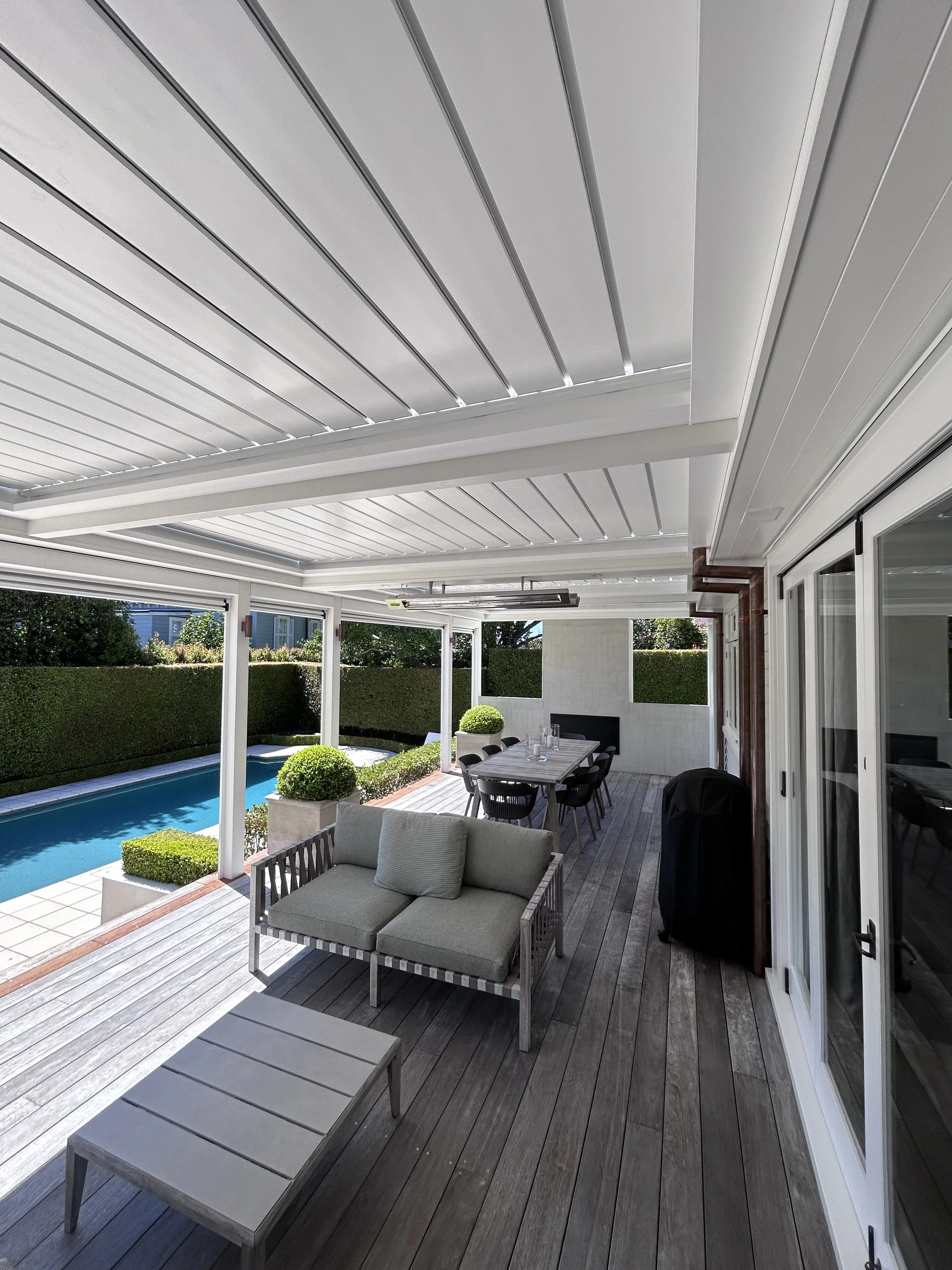 A covered backyard patio with wooden flooring, outdoor furniture including a loveseat and a dining table with chairs, surrounded by a well-maintained hedge and a swimming pool in the background.