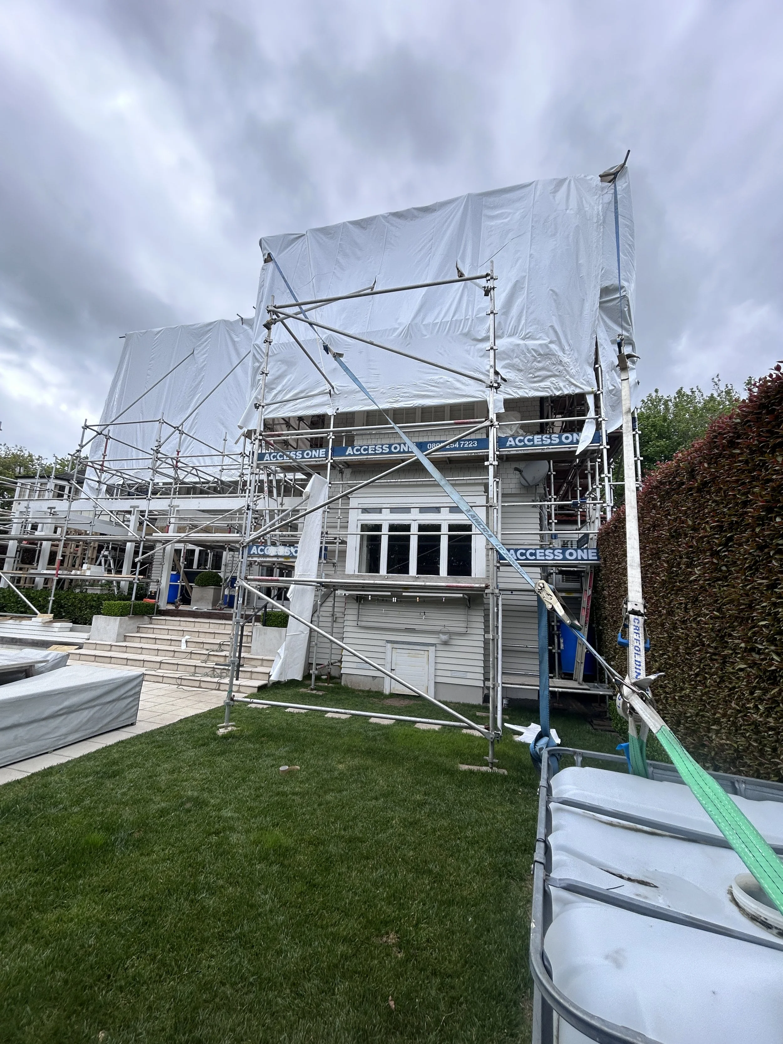 A house under renovation with scaffolding and white protective sheeting on the upper floor, with a front porch, stairs, and lush green grass in the yard.