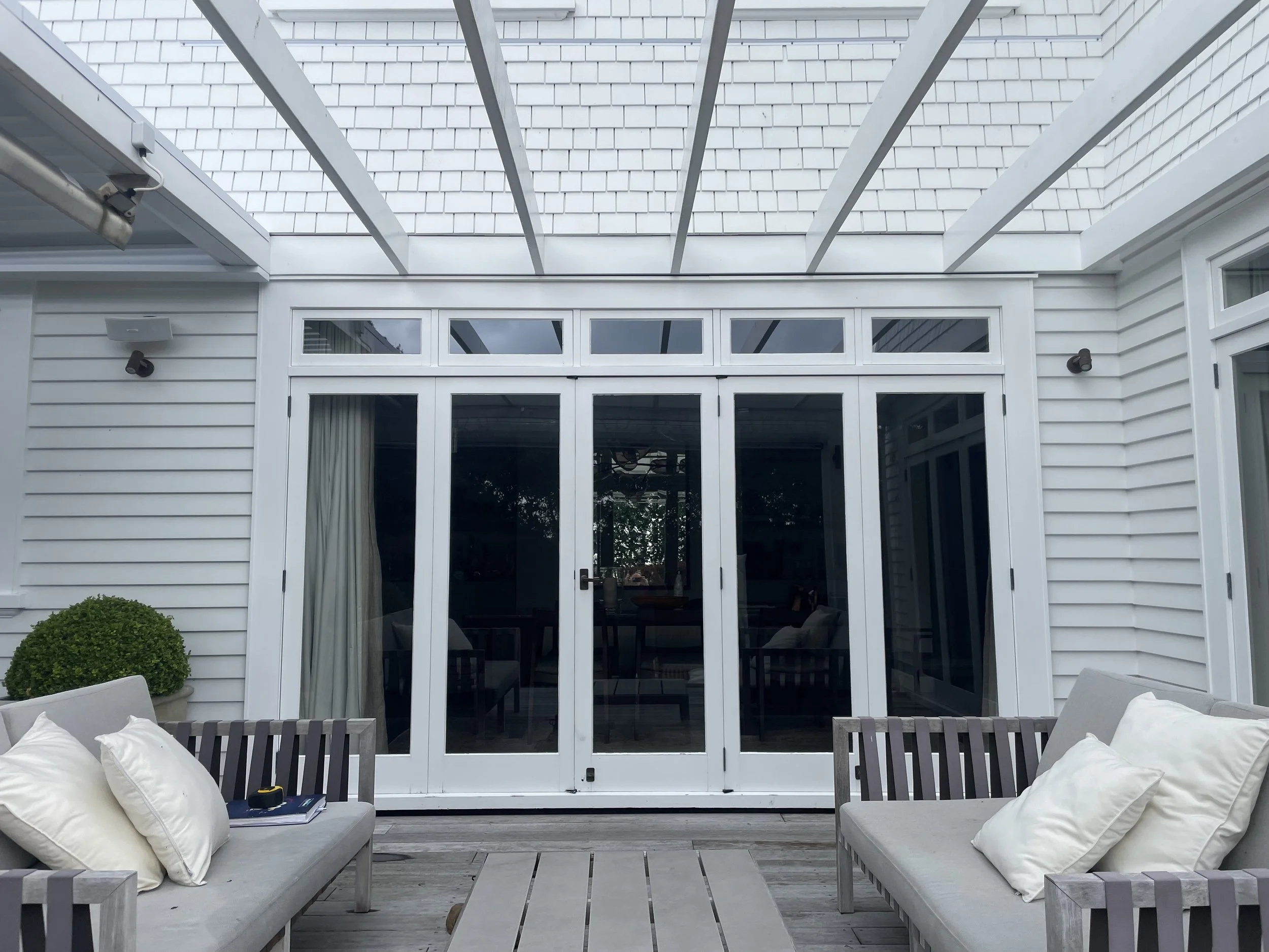 Outdoor patio area with white sofas, pillows, a table, and a large sliding glass door leading to the interior of a white house with siding.