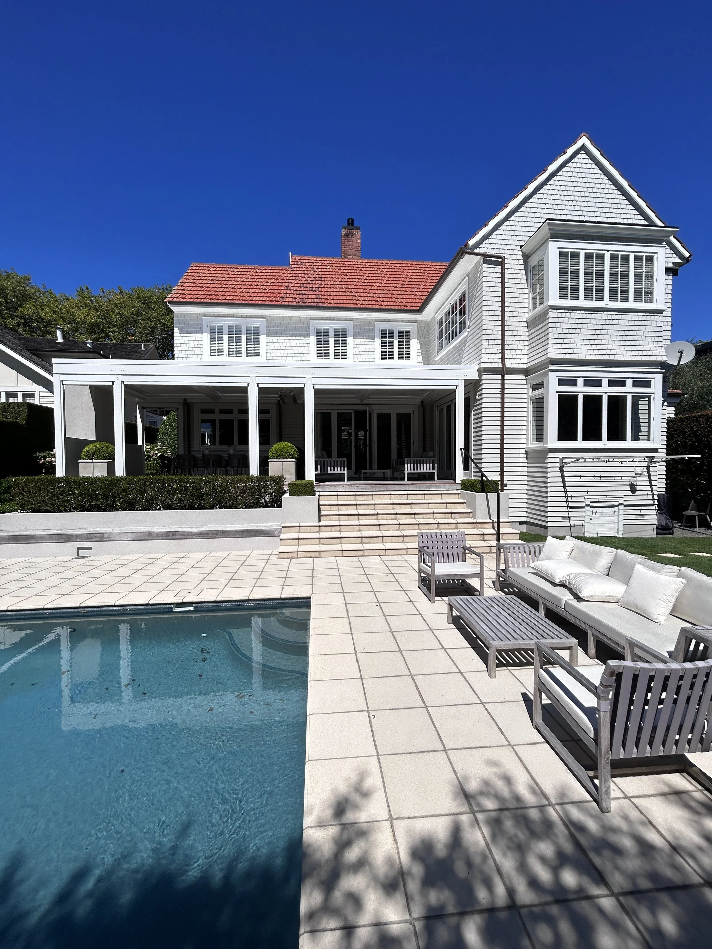 A backyard scene featuring a swimming pool, outdoor furniture including a sofa and chairs, and a white house with a red tiled roof and large windows, under a clear blue sky.