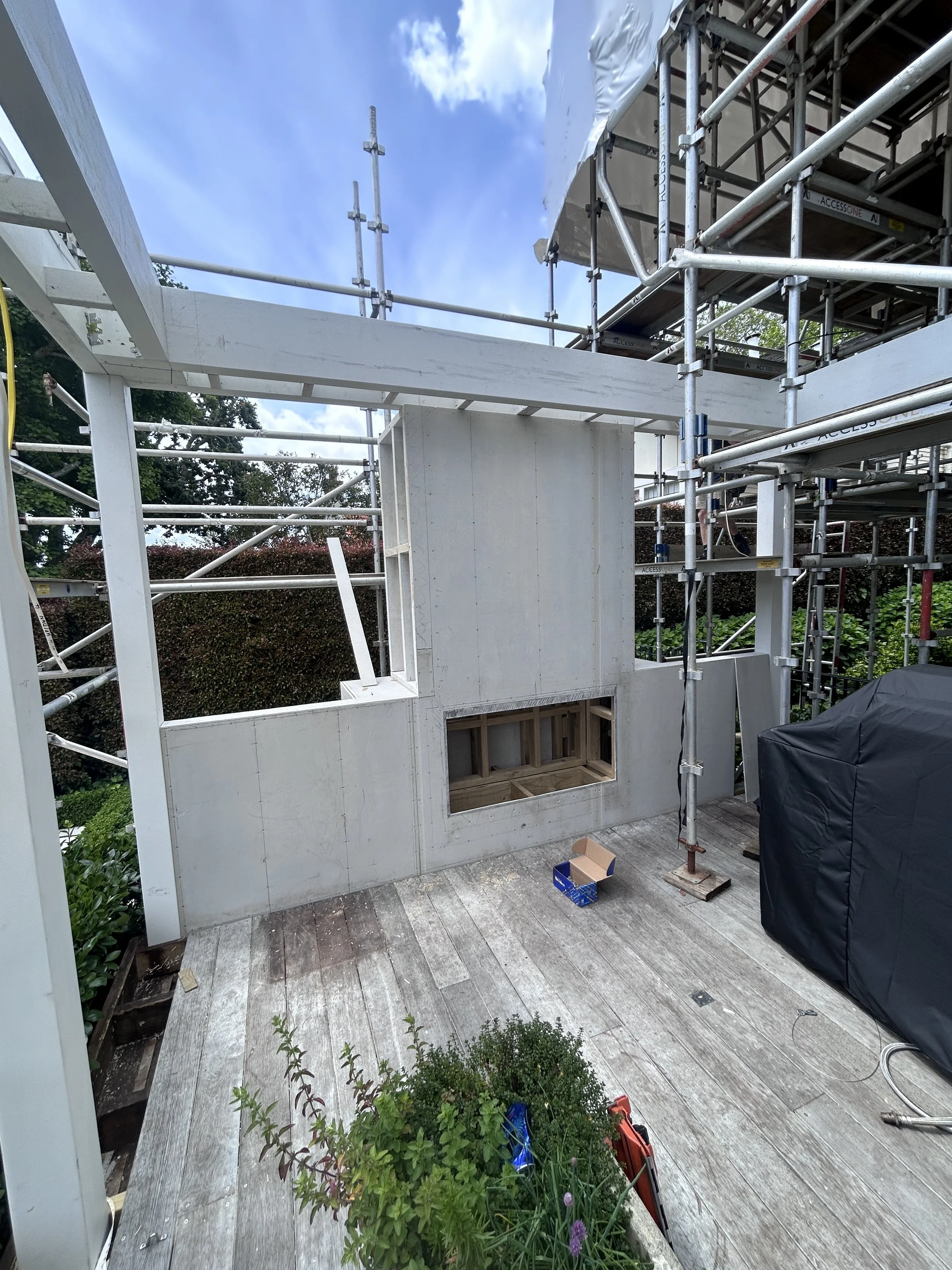 Construction site on a balcony with scaffolding and a partially built wall, potted plants, tools, and construction materials visible.