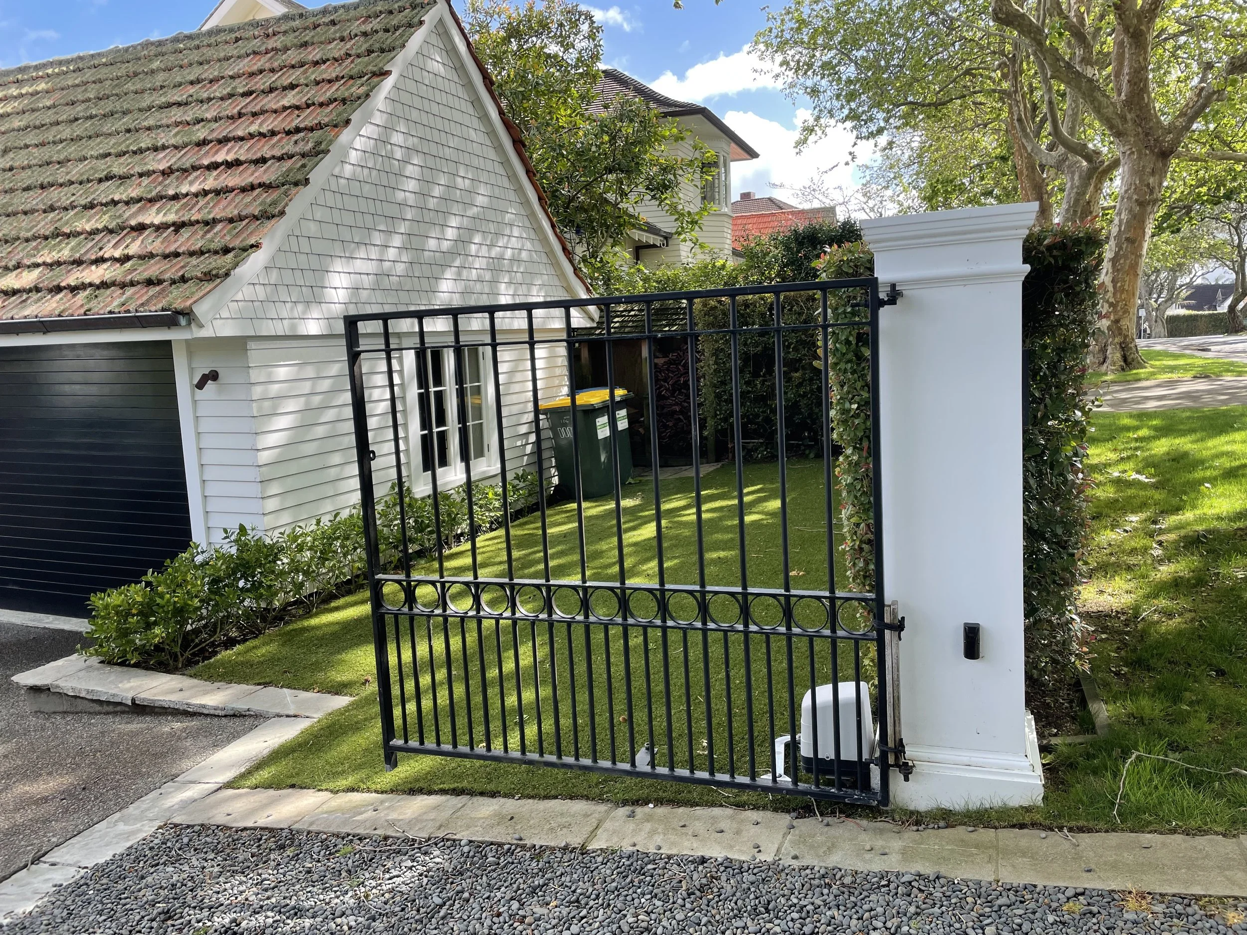 A white house with a steeply sloped, moss-covered tiled roof, visible behind a black metal gate and white pillar with a hedge and trees in the background.
