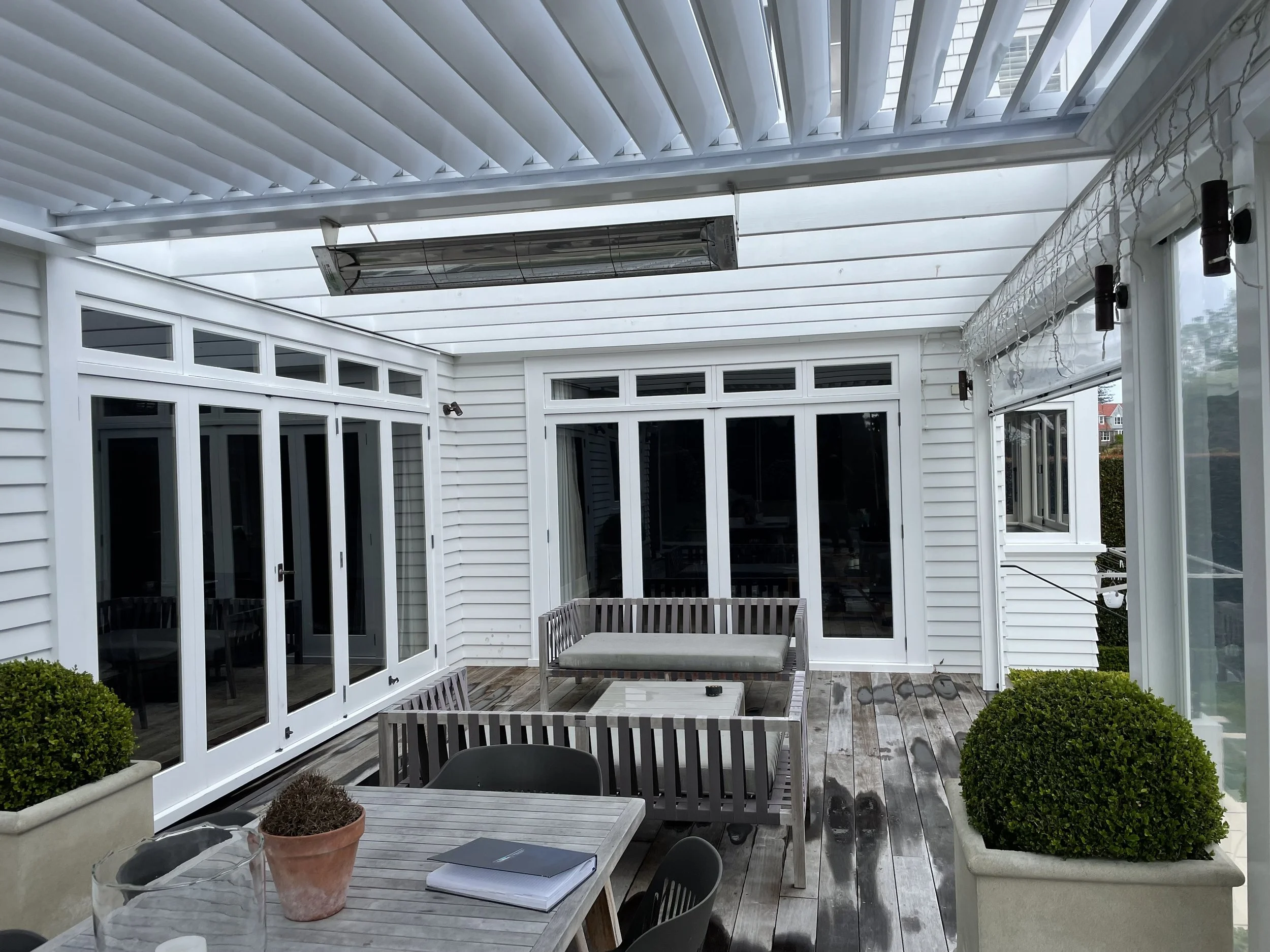 A covered outdoor patio with white siding, large glass sliding doors, and a wood deck. The patio has a white ceiling with blinds, string lights, and a heater. There's a rectangular table with a potted plant, notebooks, and glasses, surrounded by blac