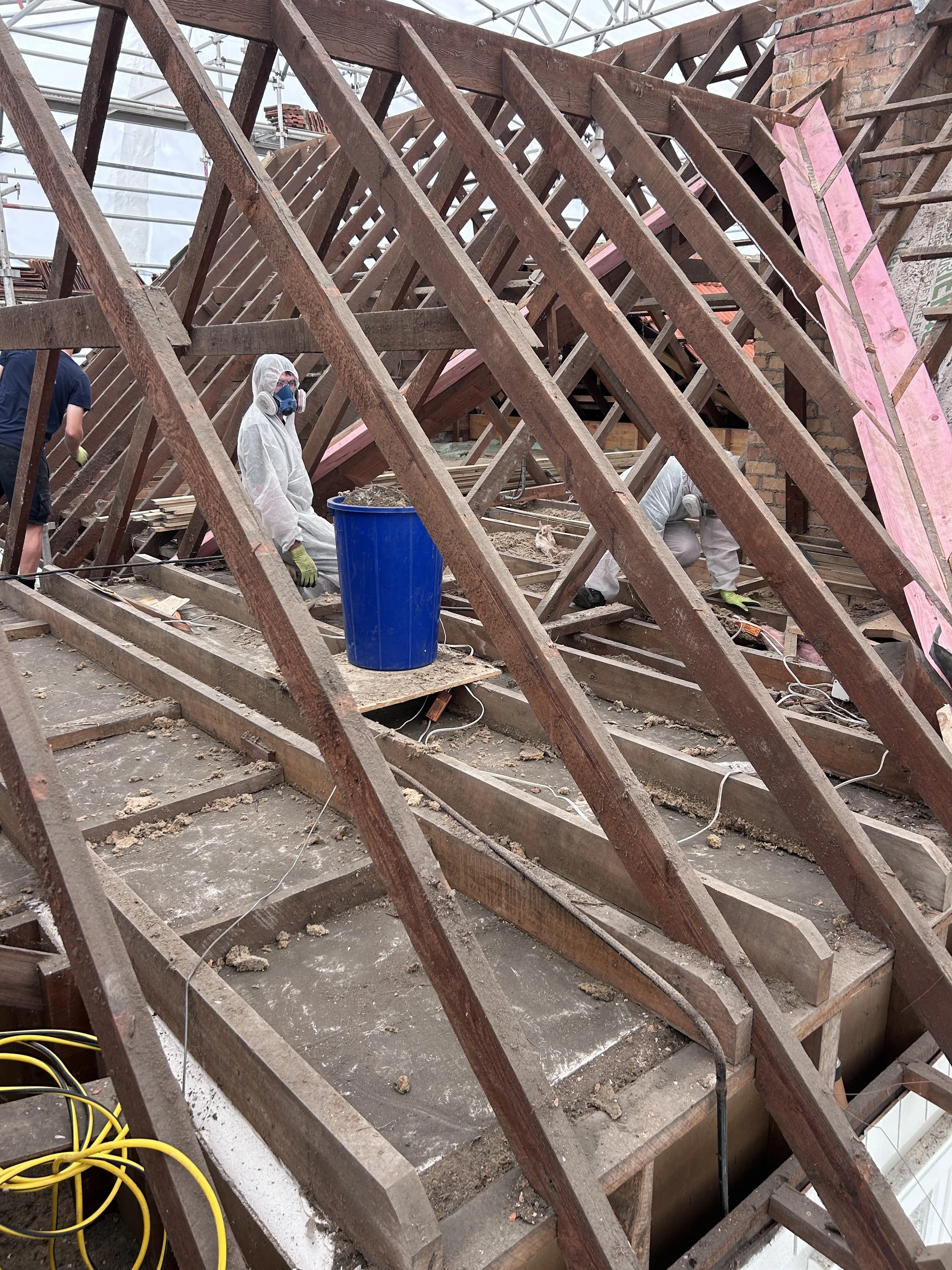 Construction workers in protective gear are working on the wooden framework of a building's roof. The image shows a wooden roof truss system, with some pink insulation panels being installed. Construction tools and yellow extension cords are visible 