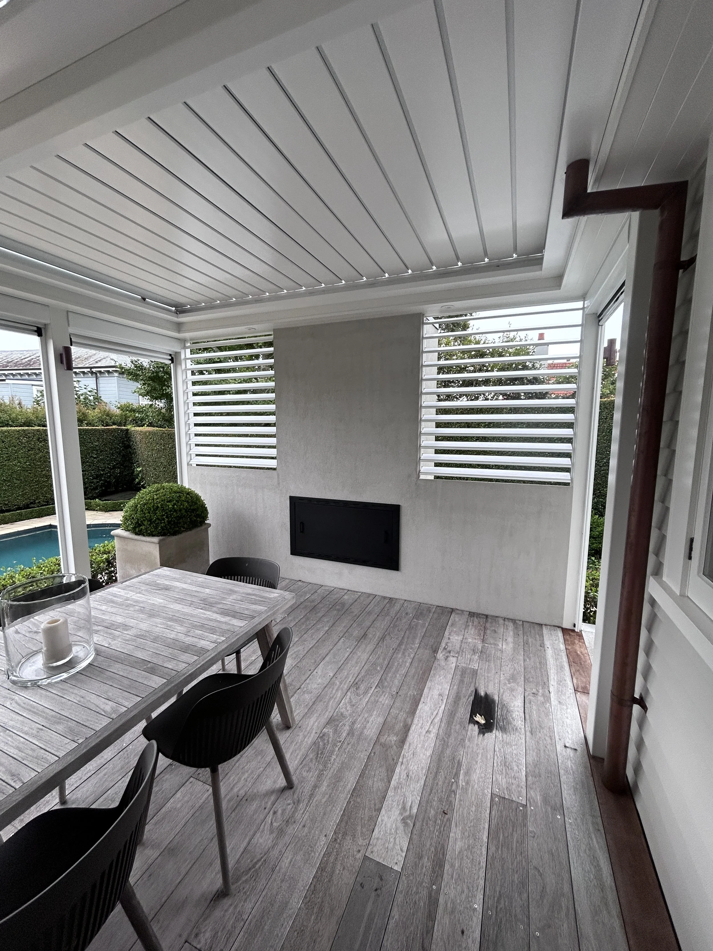 An outdoor patio area with a wooden table and black chairs, surrounded by greenery and a pool, with a white ceiling and window shutters.