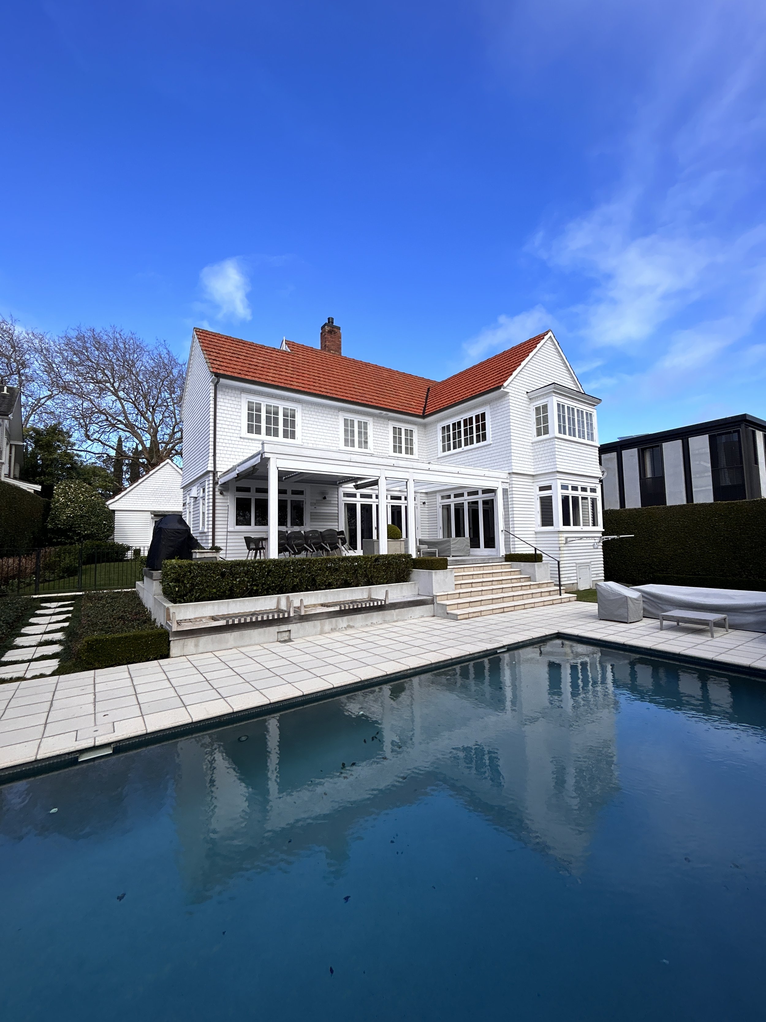 A white house with a red roof seen from the backyard, featuring a swimming pool and patio area with seating and a garden, under a partly cloudy blue sky.