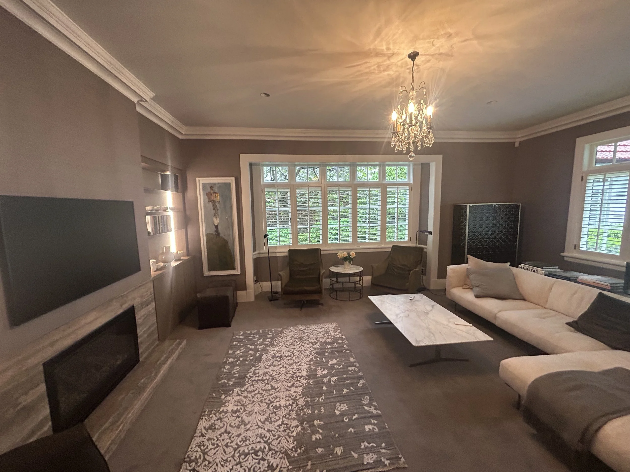A living room with a beige/white sofa, two green armchairs, a marble coffee table, a patterned rug, large windows with shutters, a chandelier, a painting, and a TV.