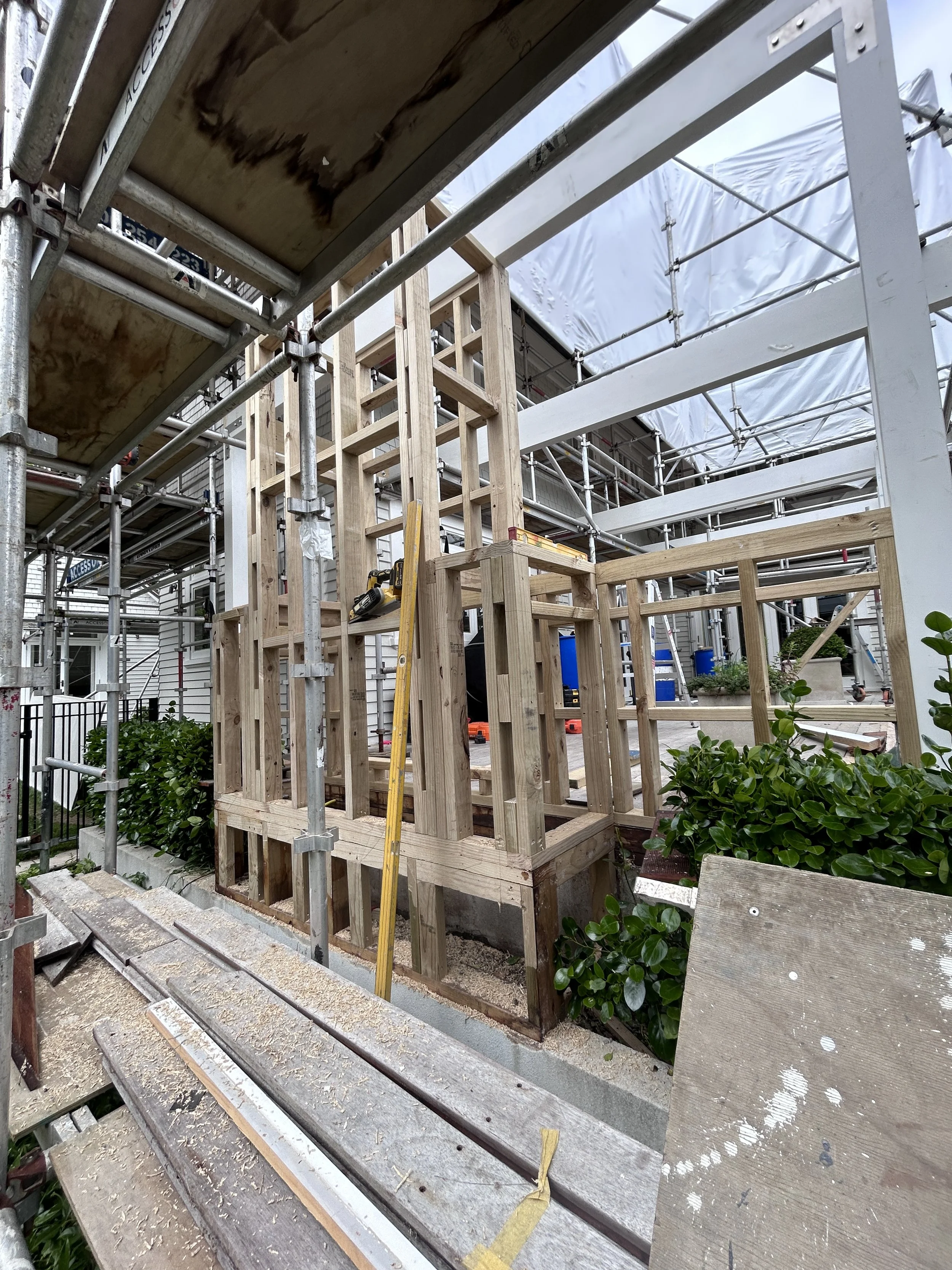Construction site with wooden framing and scaffolding around a building, with green bushes in the foreground.