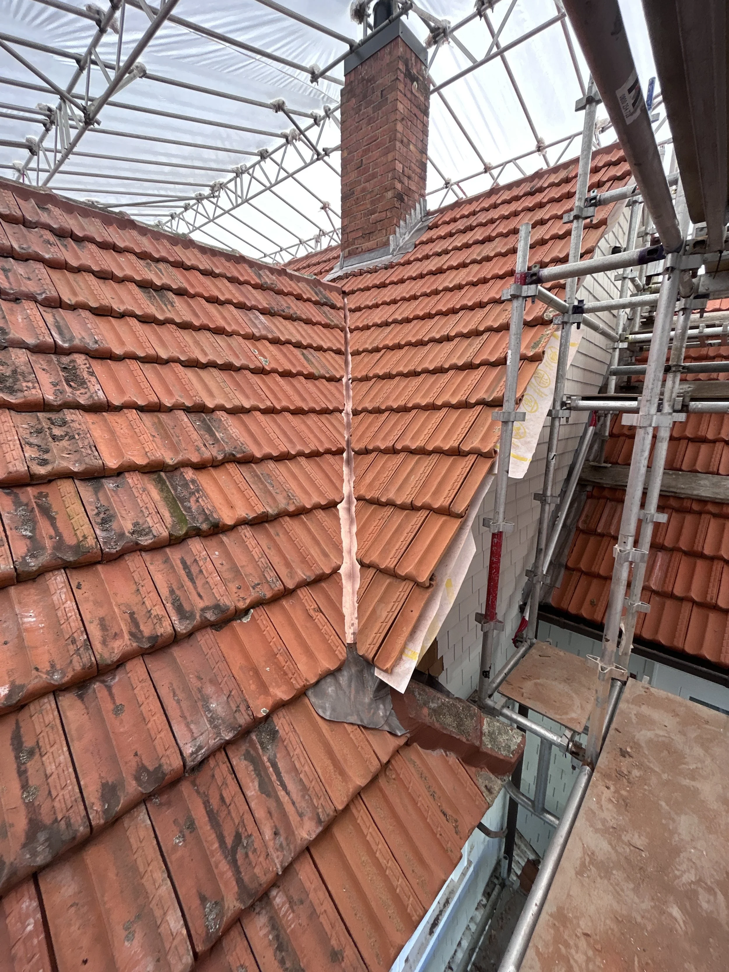 Construction scaffolding around a house with a brick chimney and a sloped roof with red tiles, some of which are weathered and discolored.