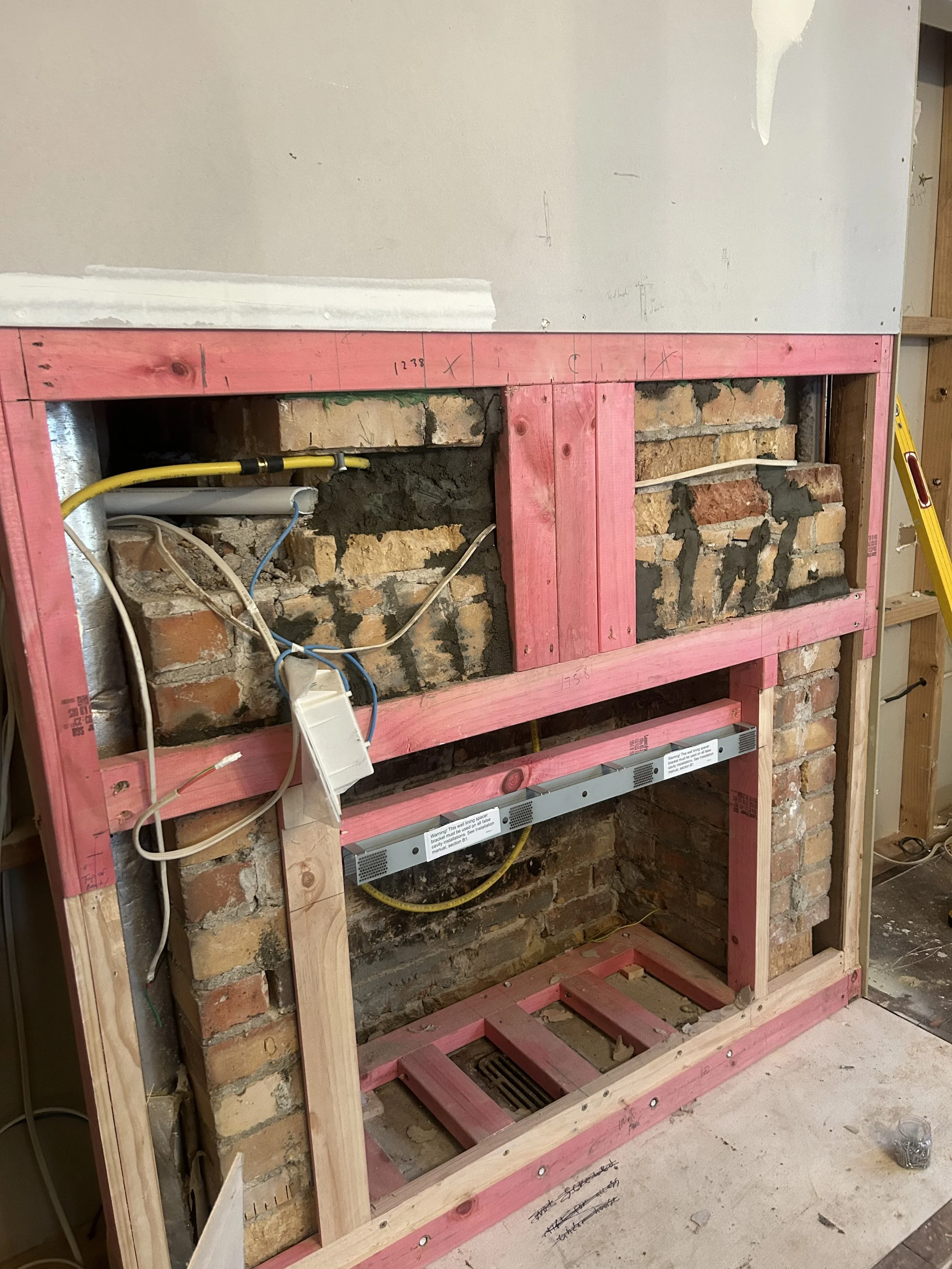 Construction of a kitchen wall with exposed brick and pink wooden framing, electrical wiring, and a level tool.