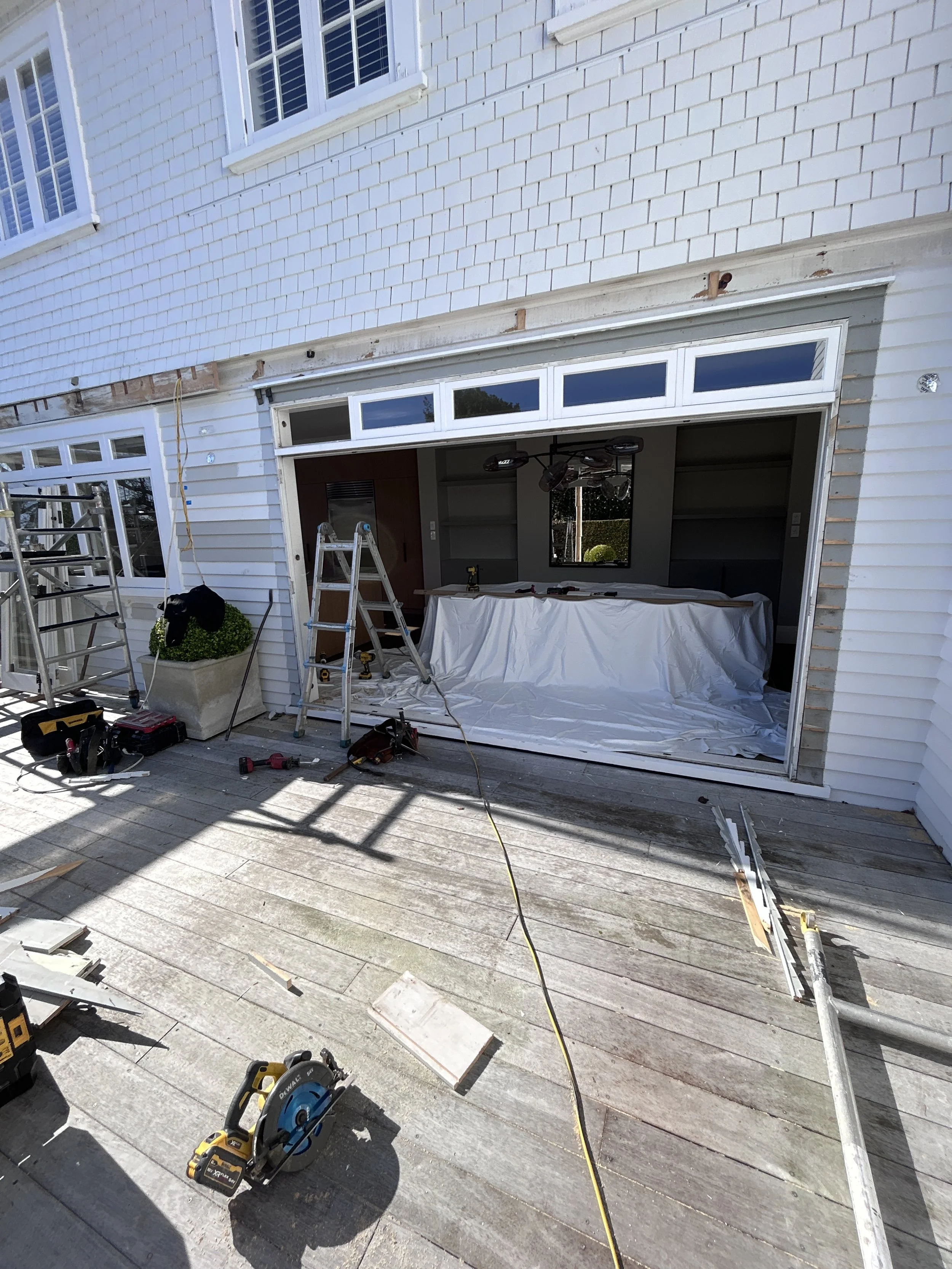 Exterior view of a house under renovation with tools and equipment on a wooden deck, including a circular saw, ladder, and construction materials, with windows and partially installed siding.