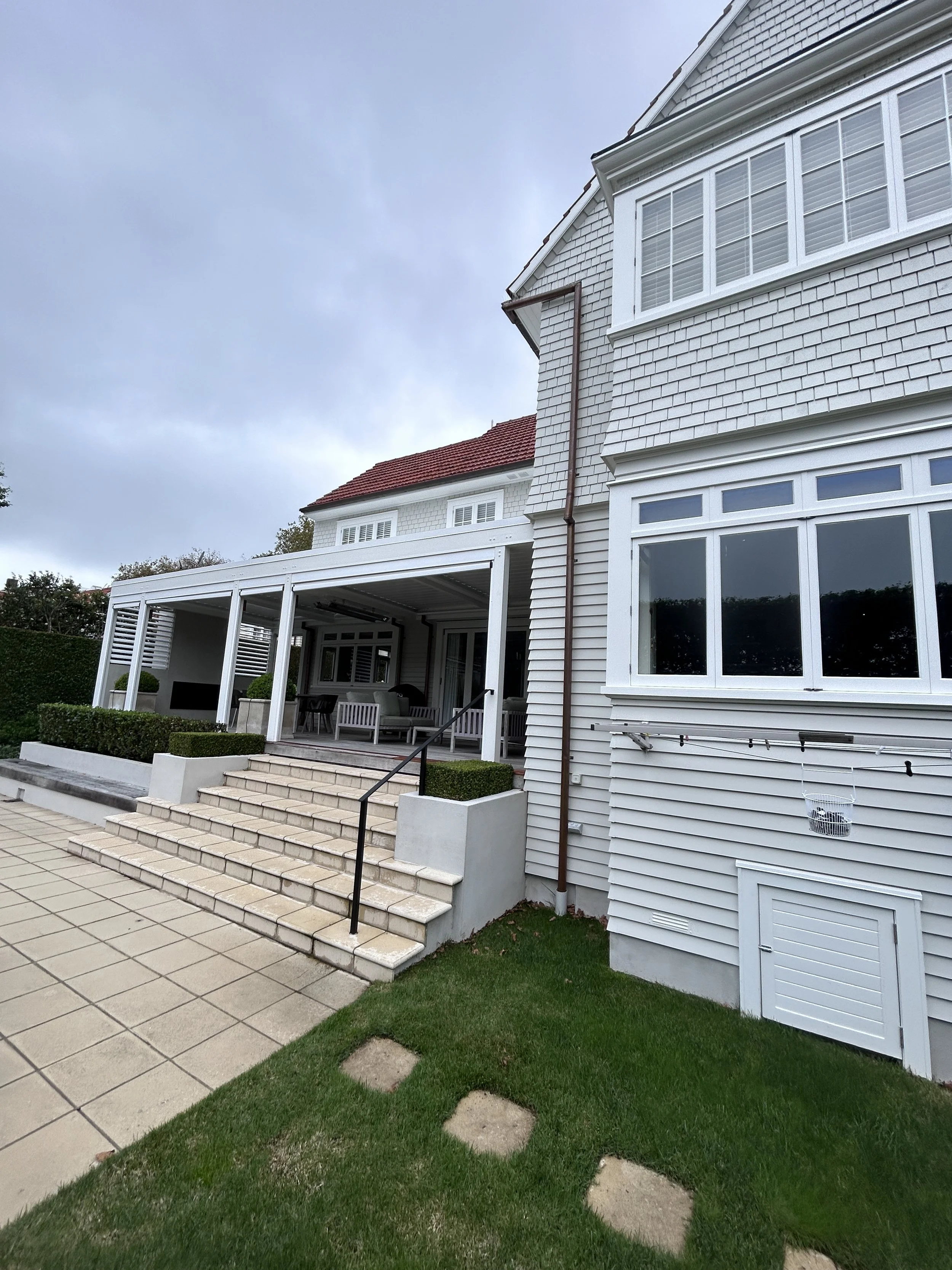 White house with large windows, red roof, and a covered patio area with outdoor furniture. Stone stairs lead to the patio, surrounded by trimmed bushes and a grassy yard, under a cloudy sky.