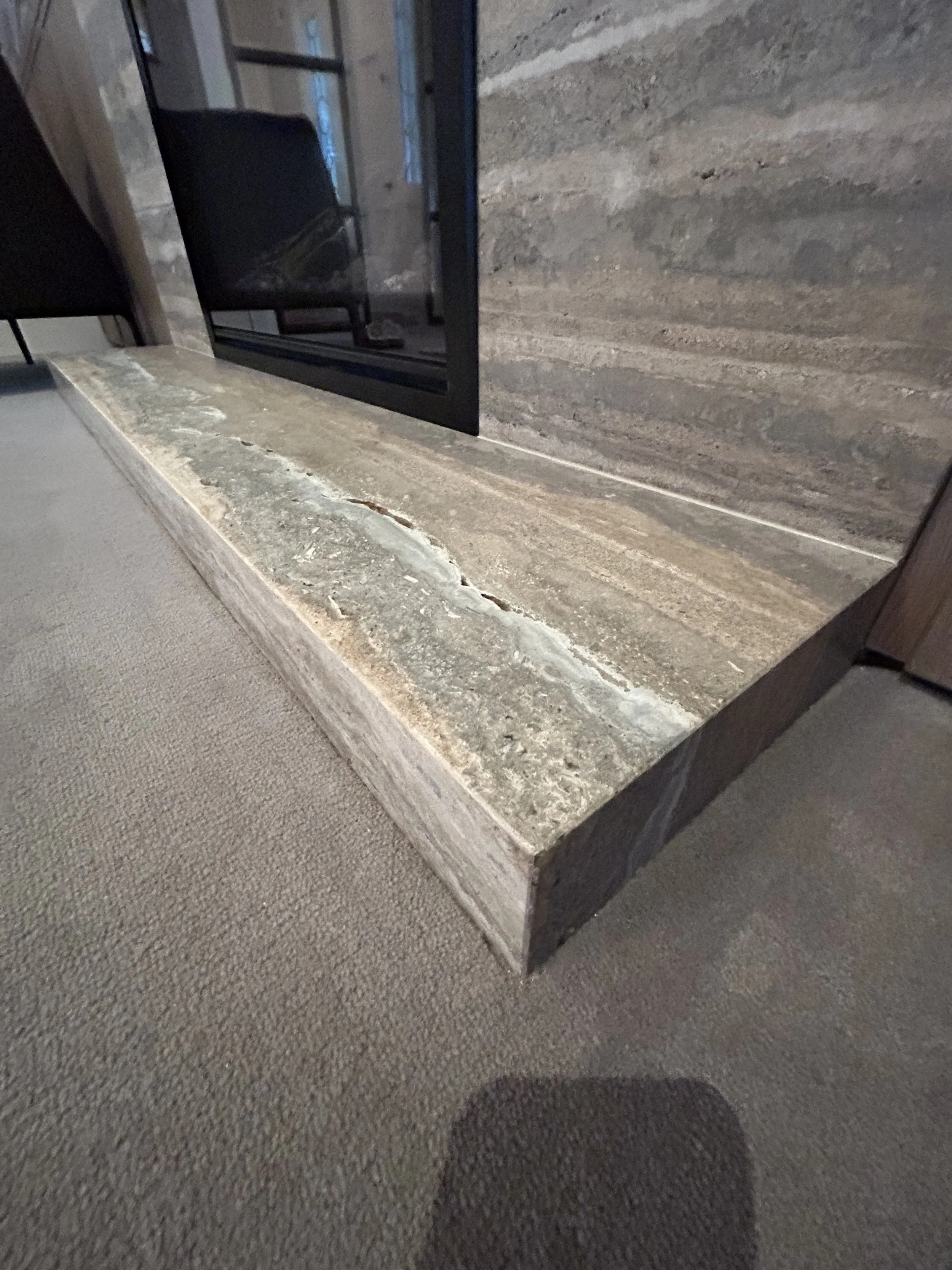 Close-up of a stone fireplace hearth with a gray and beige marbled pattern, wooden mantel, and a dark-colored firebox opening.