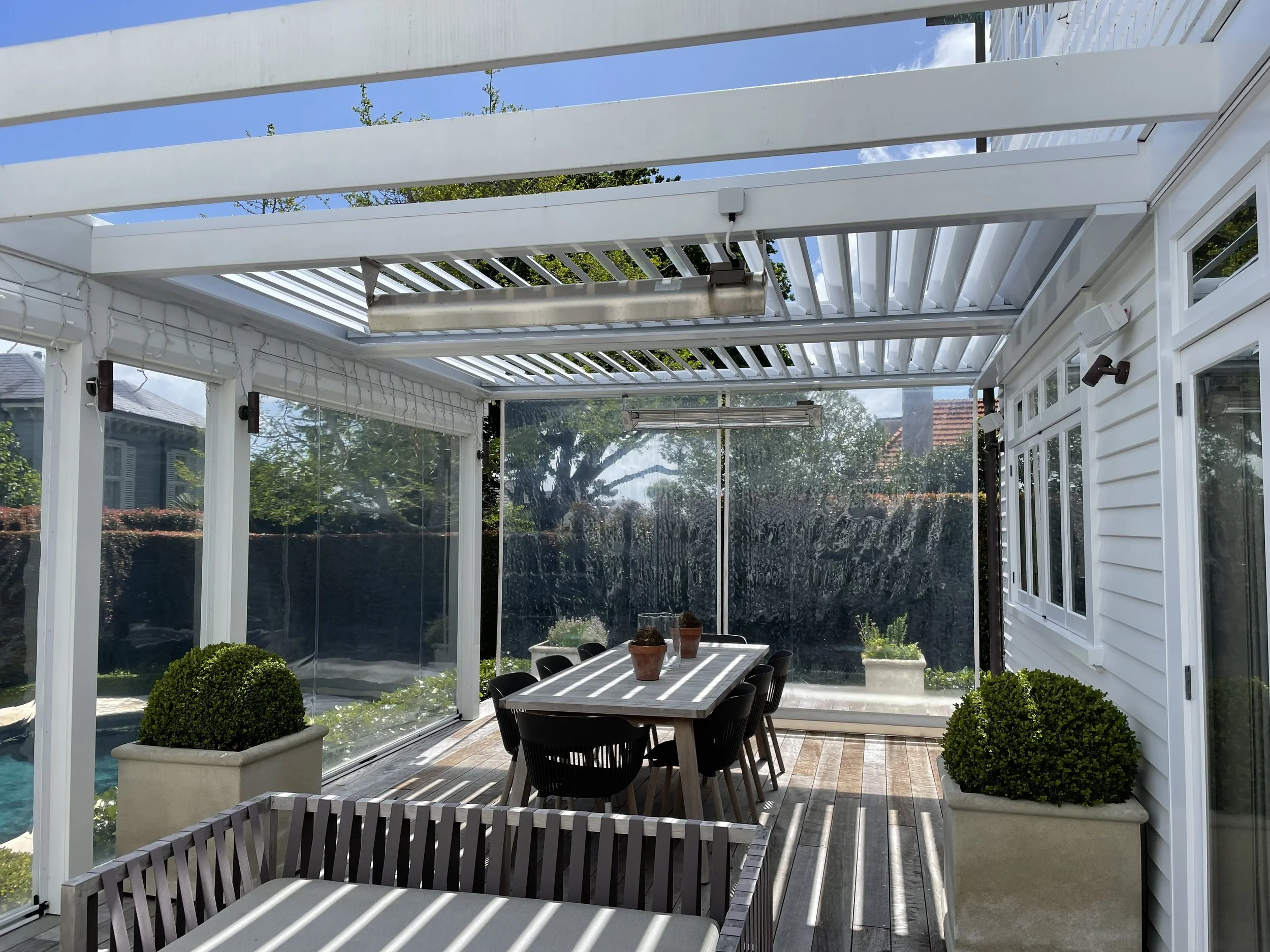 A sunroom with glass walls and ceiling, white wooden beams, a dining table with six chairs, potted plants, and outdoor scenery in the background.