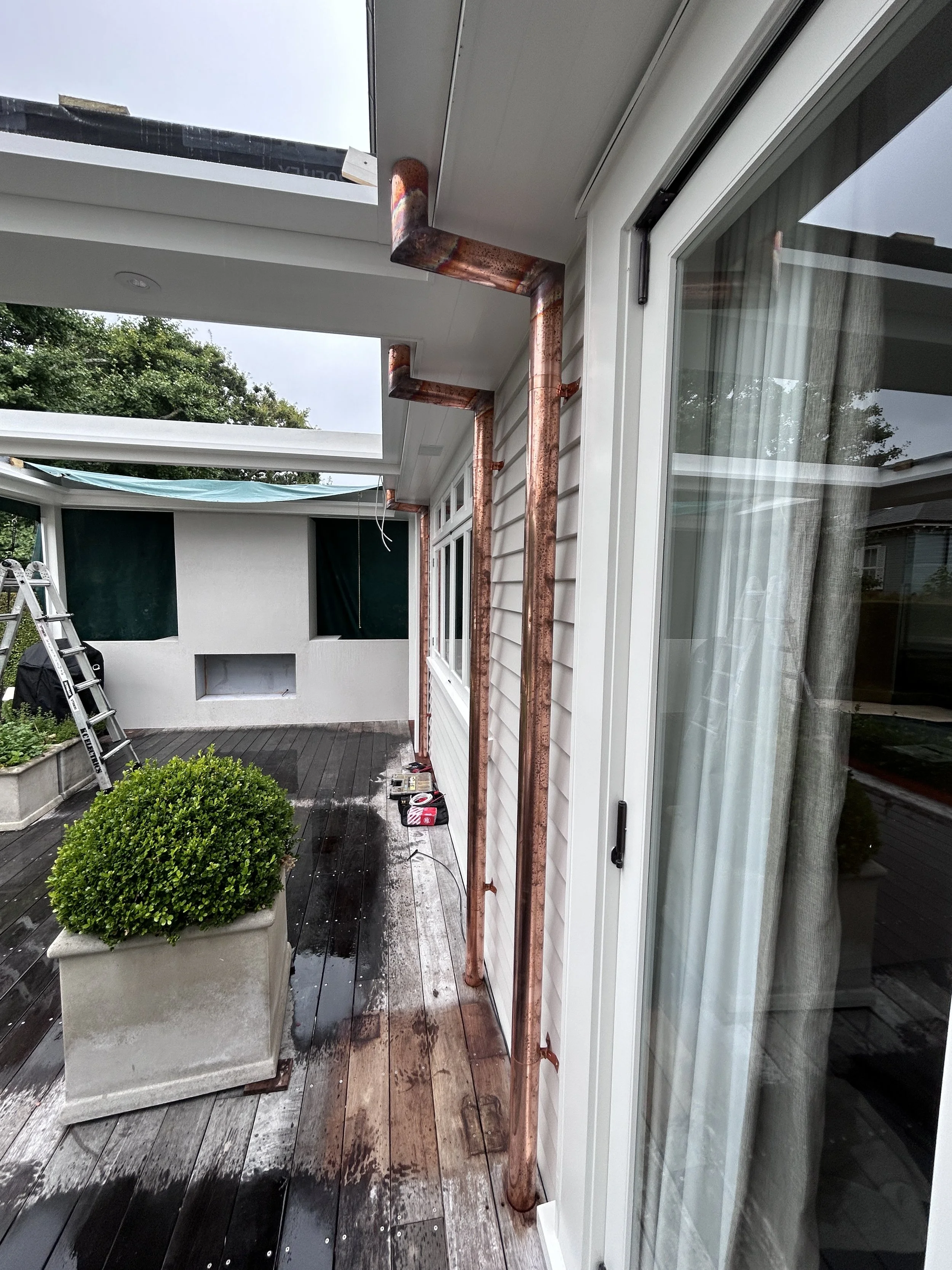 Exterior porch with a potted green shrub, a stepladder, and copper pipes installed along the wall under a cloudy sky.
