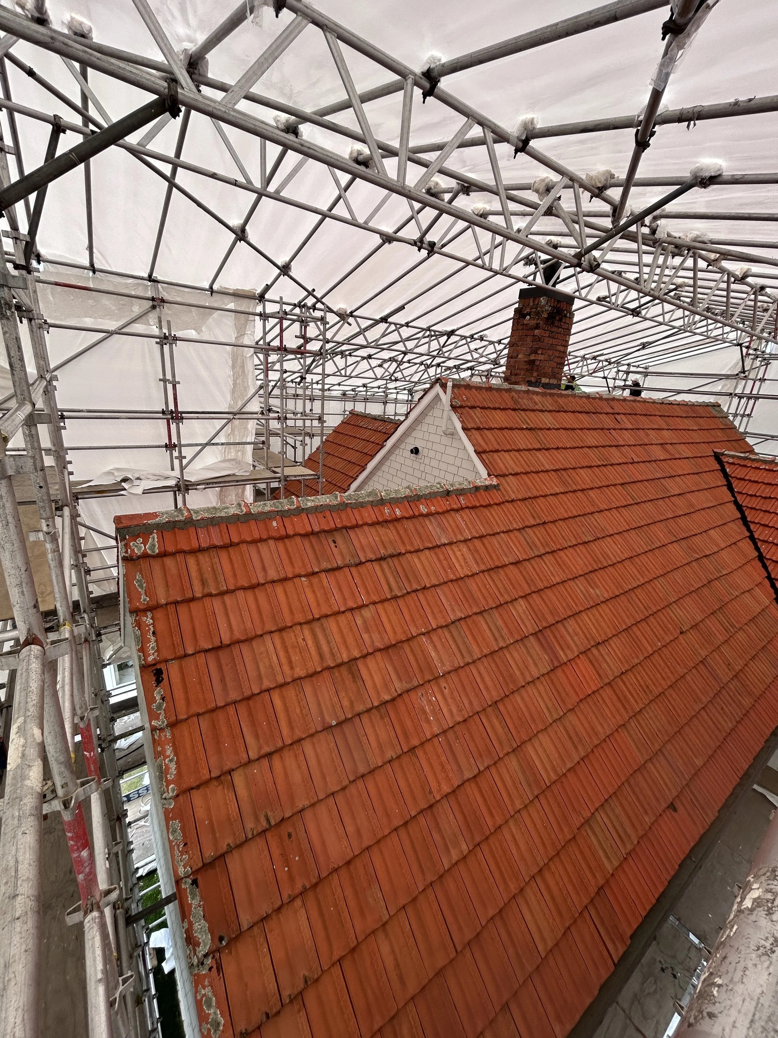 Scaffolding surrounds a house with a red tiled roof, white brick exterior, and brick chimney, indicating construction or renovation work.