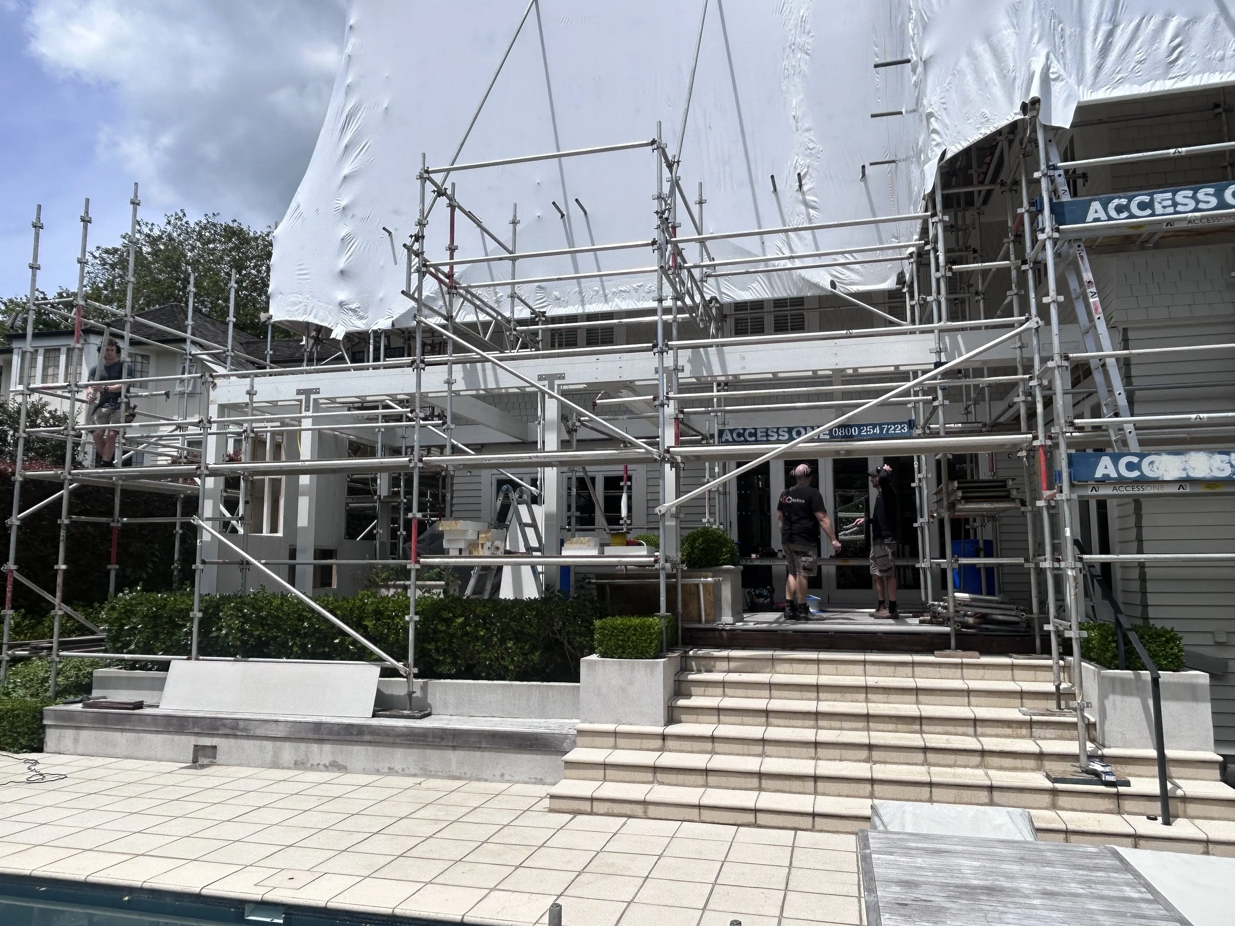 Scaffolding set up around a house with crew members working, in front of a stairway and a small garden.