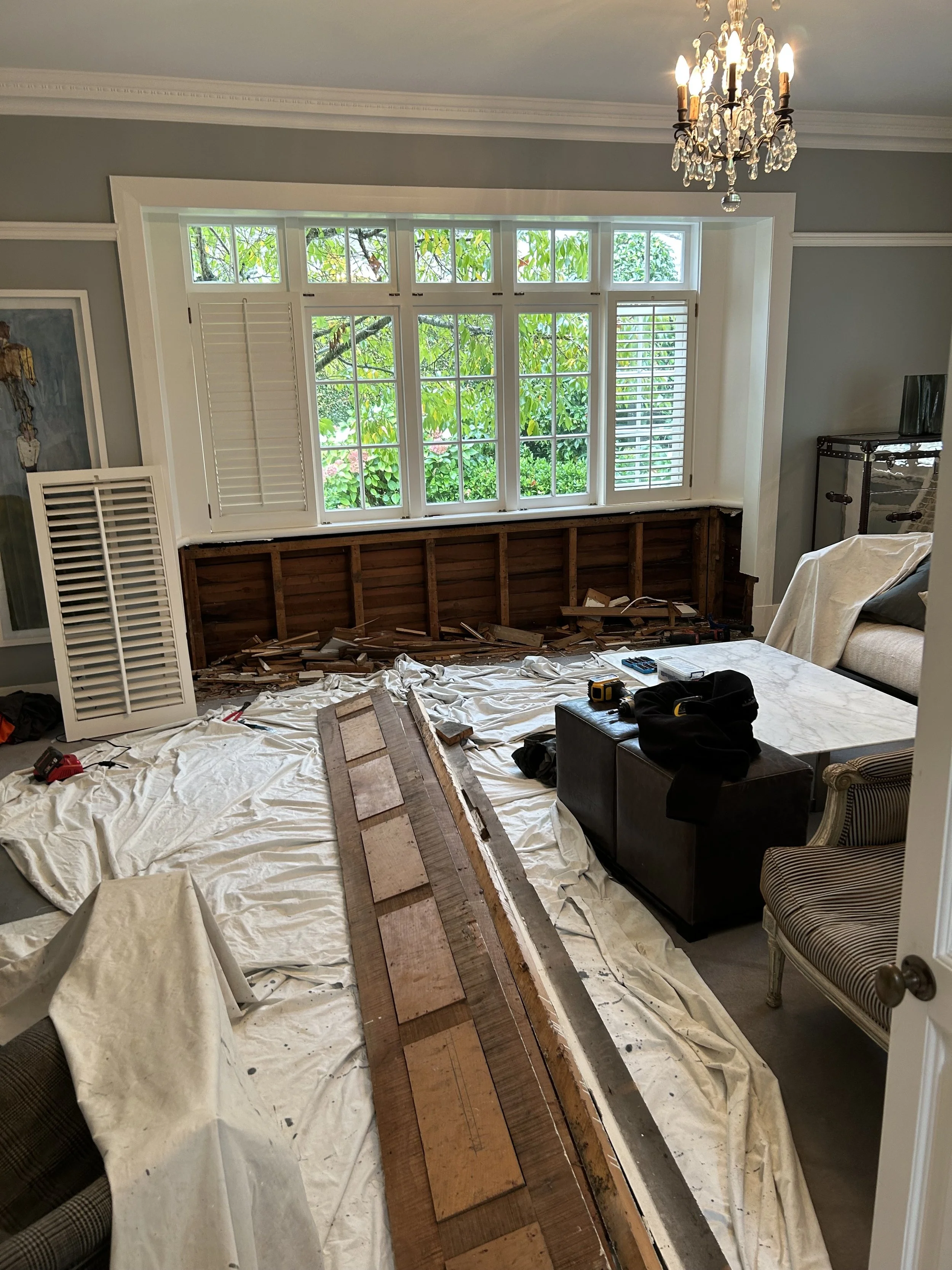 A living room in the process of renovation with a large window, some furniture, and tools on the floor. The lower part of a wall has been removed, exposing wooden studs. A chandelier hangs from the ceiling.