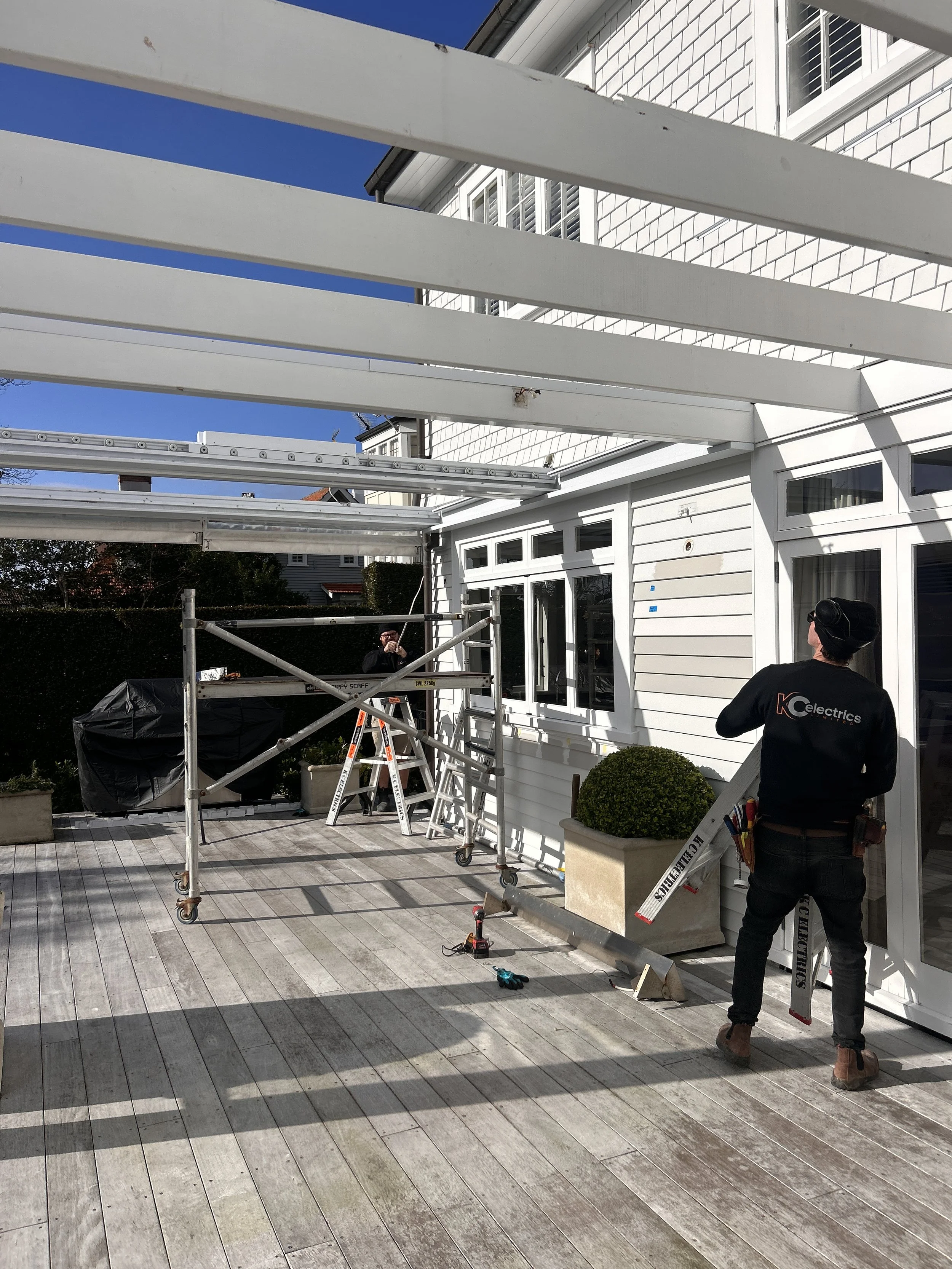 Workers installing a pergola or awning frame on a patio outside a white house with large windows, construction tools and scaffolding present
