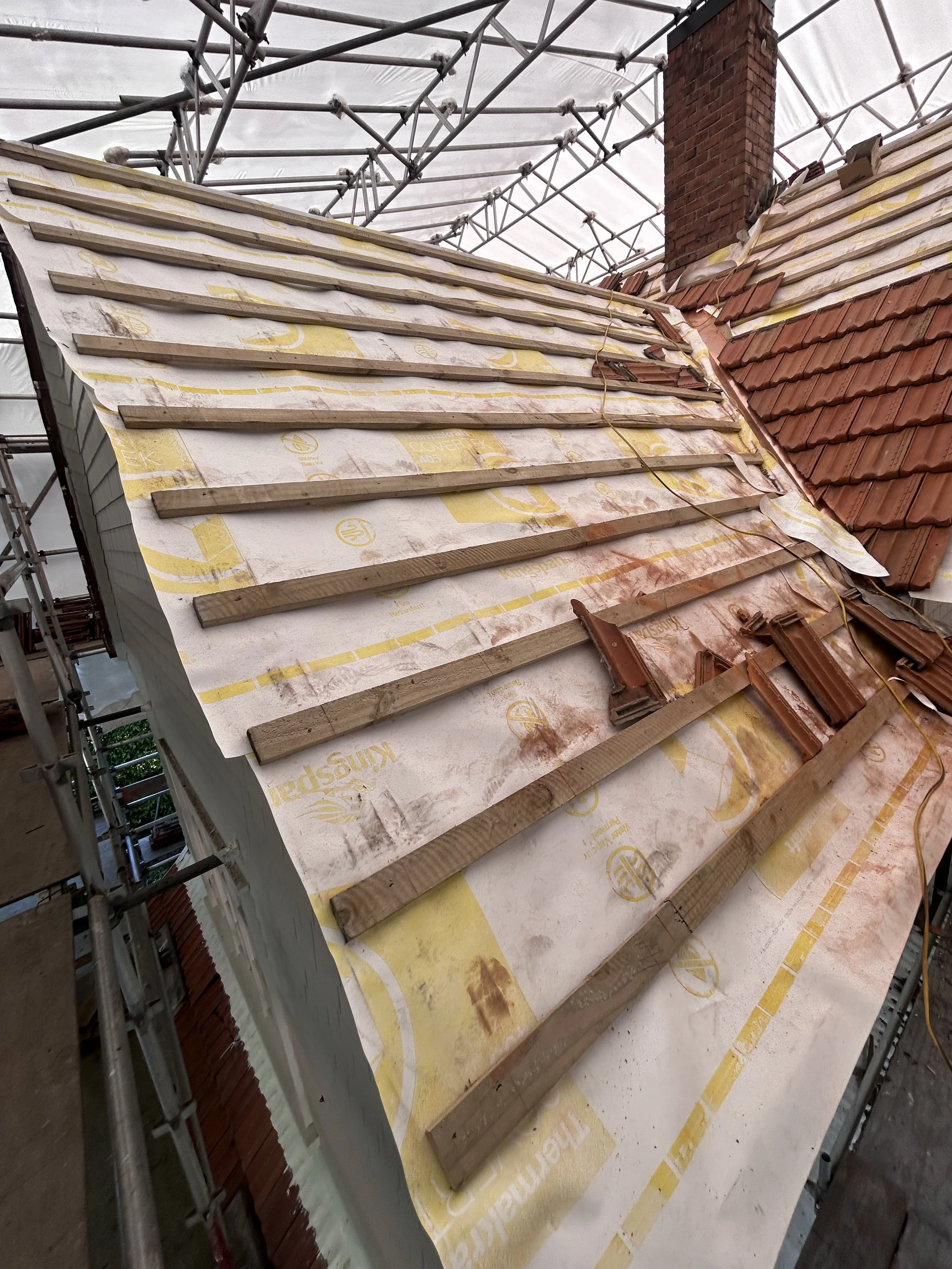 Construction site showing a roof being repaired with wooden battens, tiles, and scaffolding, with a brick chimney in the background.