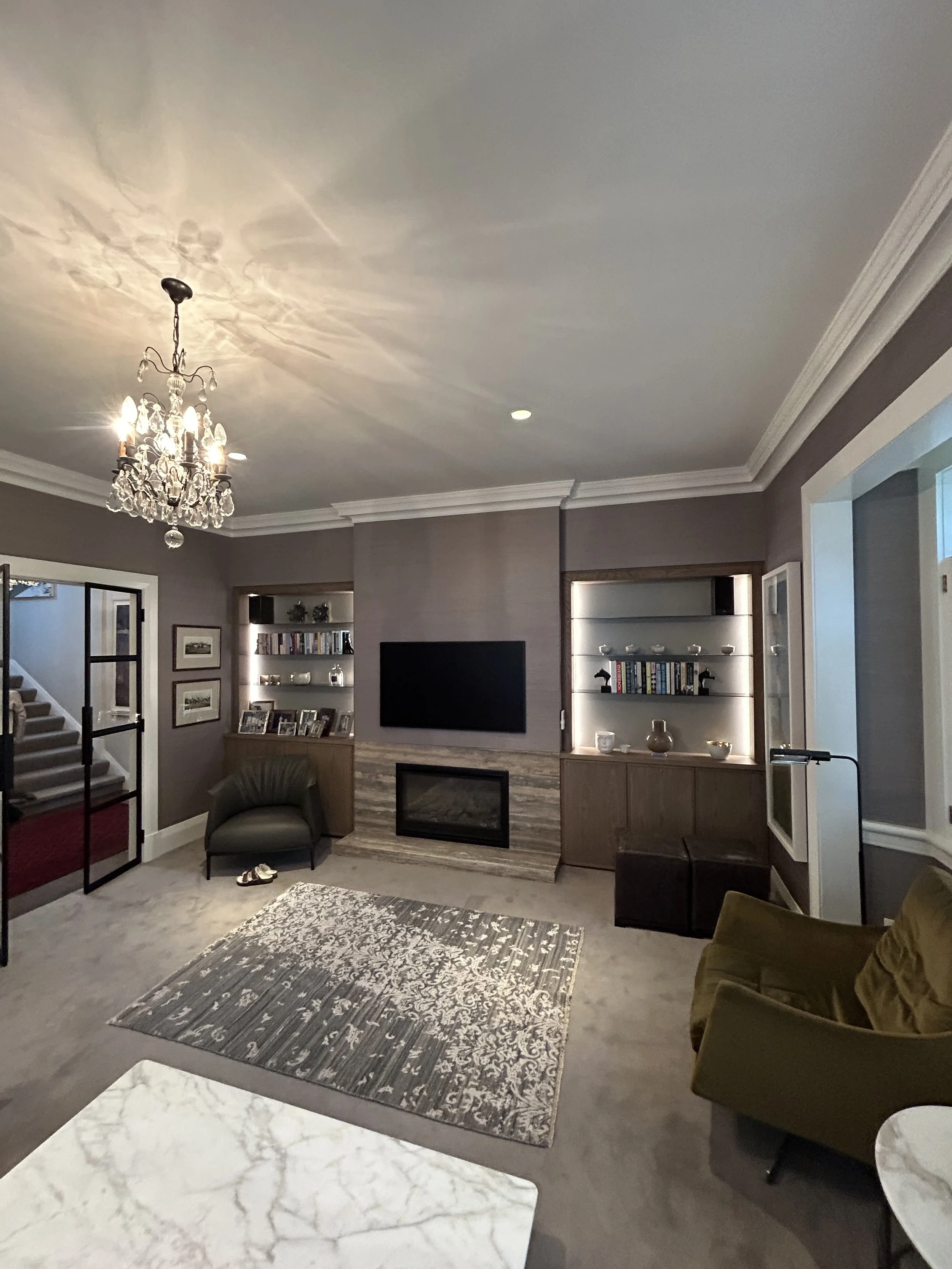 Living room with gray walls, white crown molding, a chandelier, a fireplace with a marble surround, a wall-mounted flat-screen TV, bookshelves with decorative items, a patterned area rug, and a green armchair.