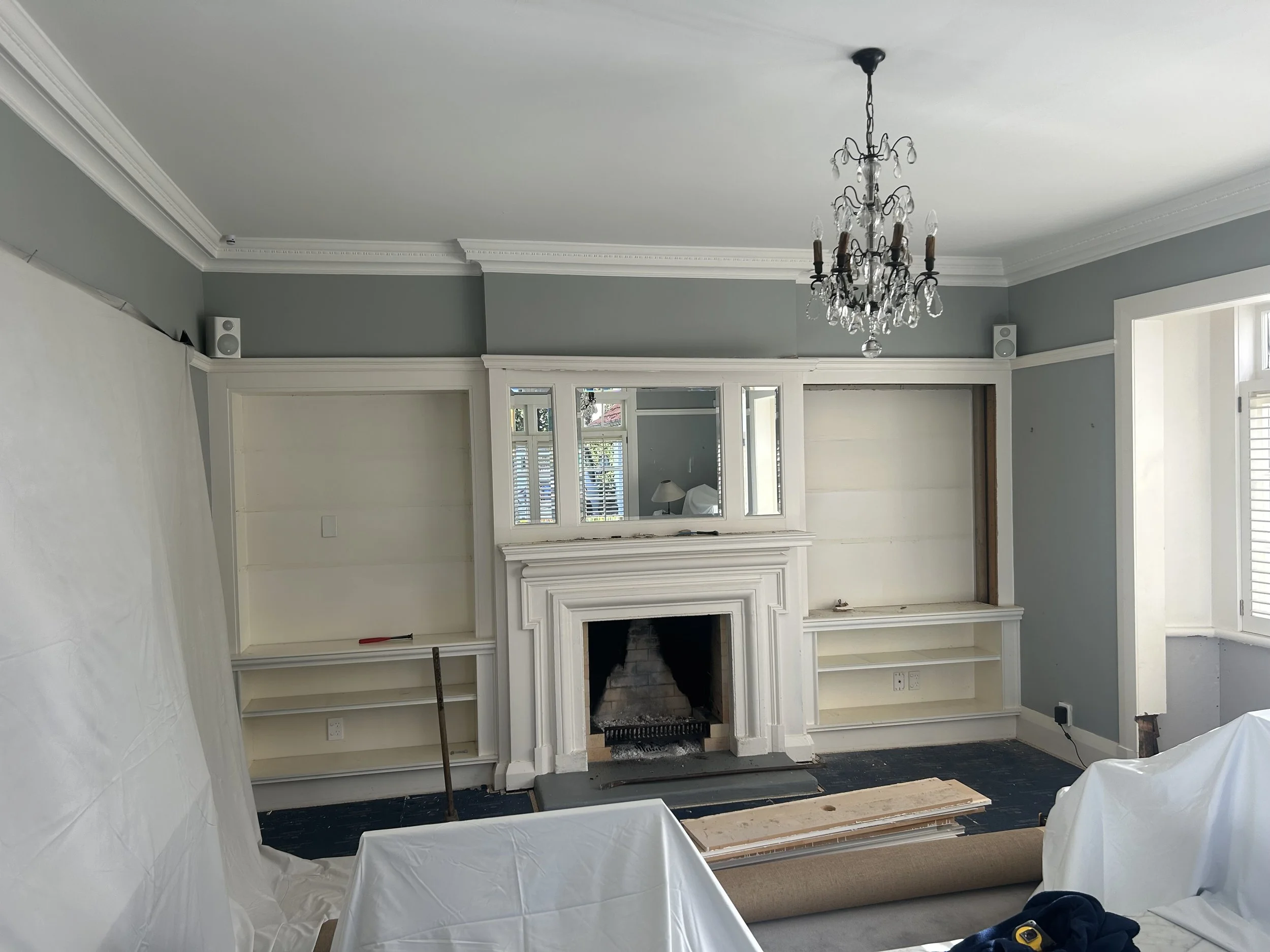 Living room with white built-in shelves and fireplace, small chandelier hanging from ceiling, some construction or renovation work in progress