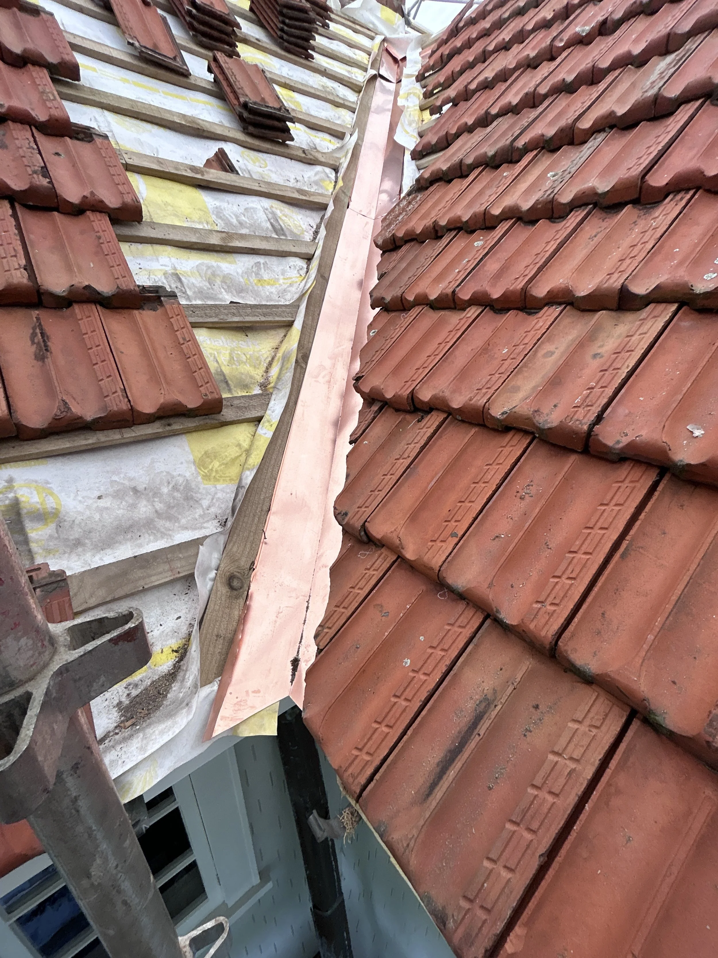 A rooftop with red clay tiles, some of which are being replaced or repaired, with a wooden frame and insulation underneath. Part of a building's exterior and windows are visible in the background.