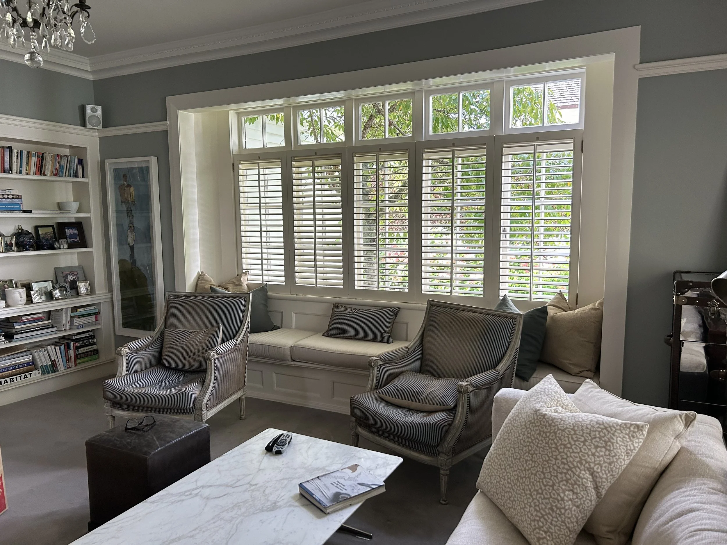 Living room with a large window with white shutters, a bookshelf filled with books and photos, and several armchairs and sofas with beige and striped cushions.