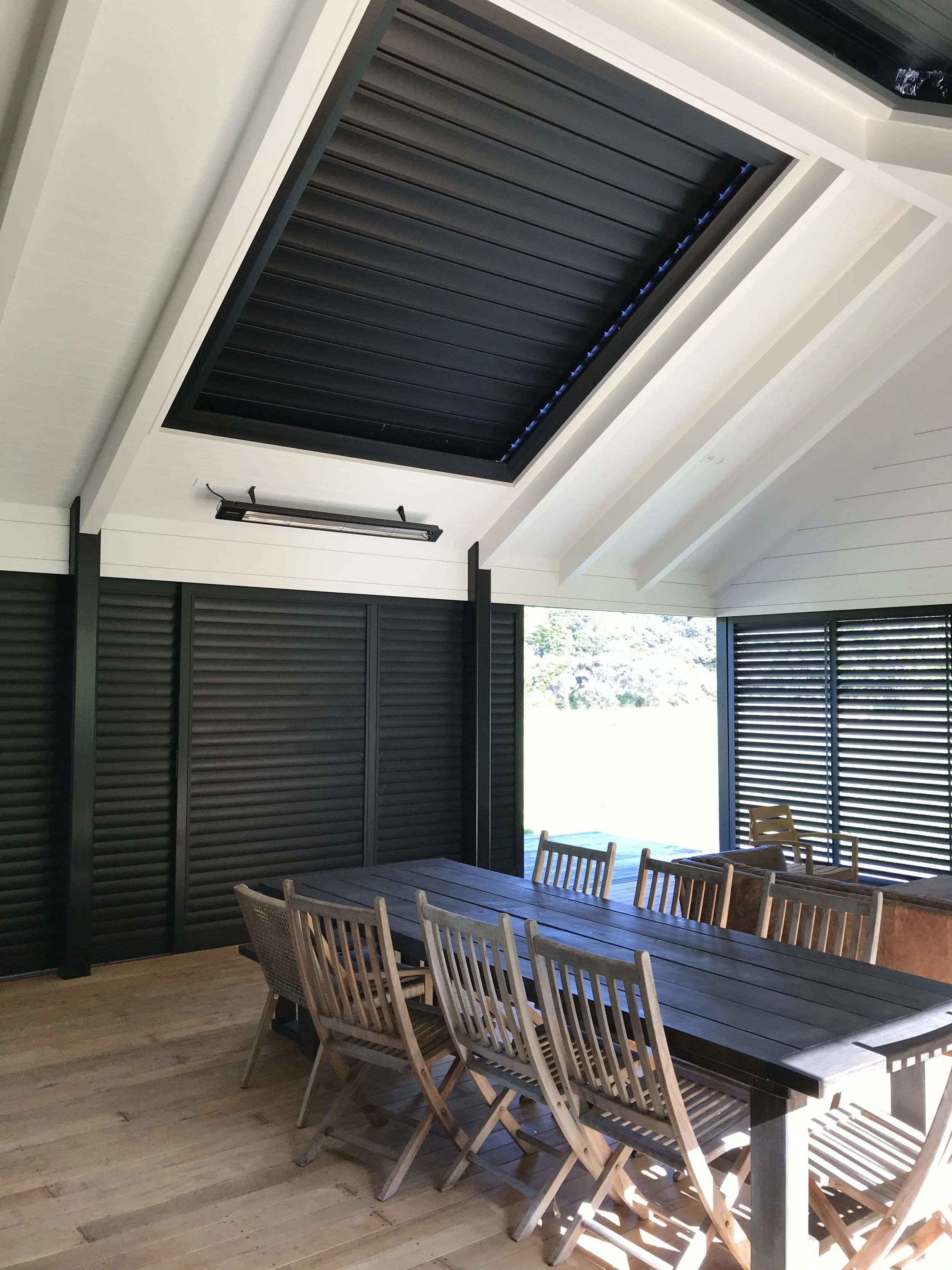 An indoor dining area with a long wooden table surrounded by wooden chairs and a large window with black shutters letting in natural light.