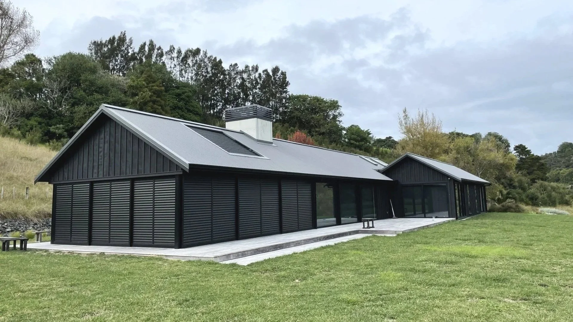 A modern black house with a metal roof and large glass windows, situated on a grassy lawn with a backdrop of trees and hills under a cloudy sky.