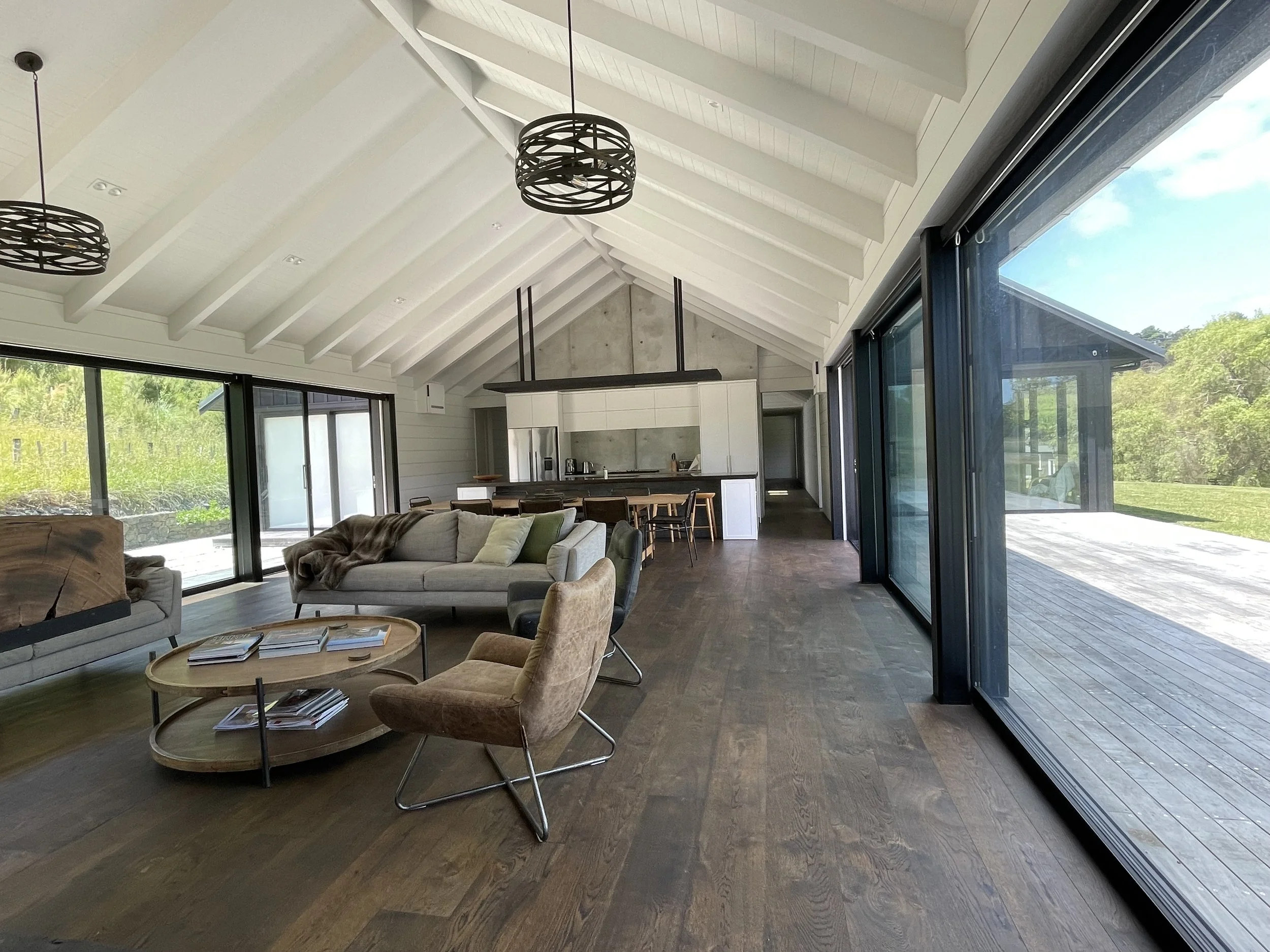 Open-concept living area with high vaulted white ceiling, exposed beams, large sliding glass doors, and modern furnishings including a beige sofa, a round coffee table with magazines, and a dining table with chairs, overlooking a lush green outdoor l