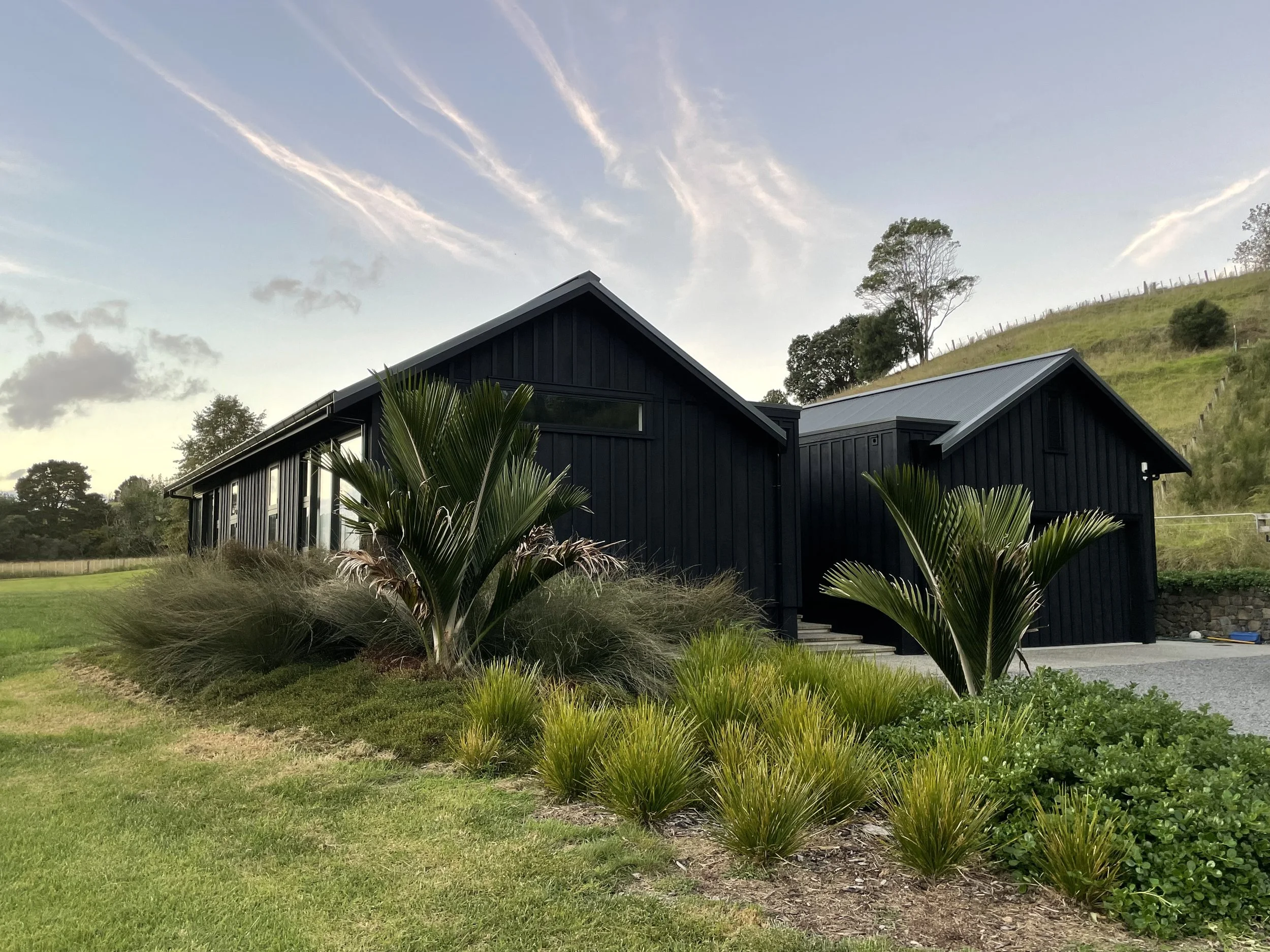 A black modern house with large windows, surrounded by green plants and grass, with a hillside and trees in the background, under a partly cloudy sky.