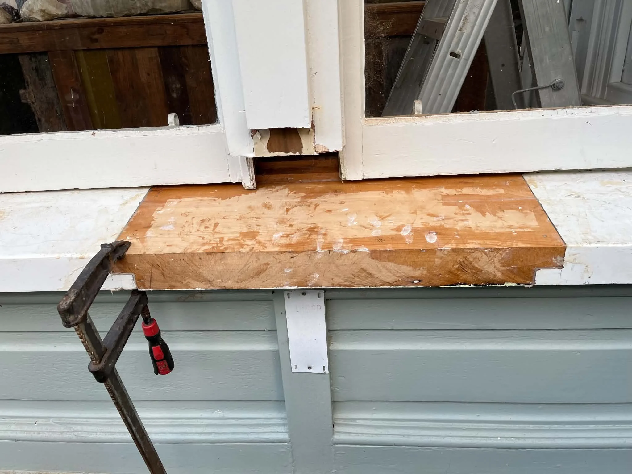 A work area with a small portable saw horse and a plastic clamp in front of a partially cut wooden frame with white paint, with some tools and ladders visible in the background.